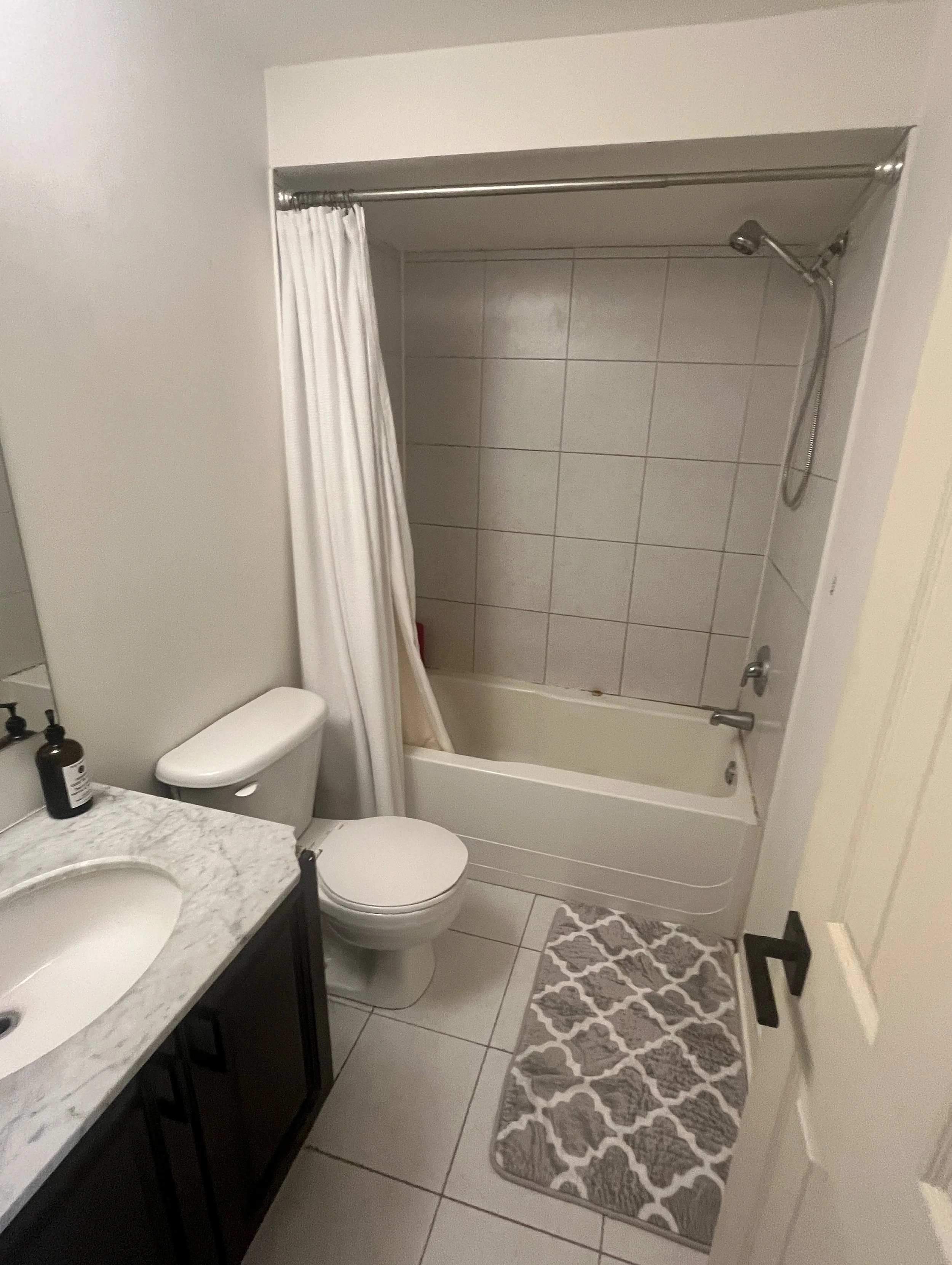 Before photo of a dated bathroom with a white tub-shower combo, small white floor tile, marble-top vanity, and grey patterned bath mat, ahead of a full renovation