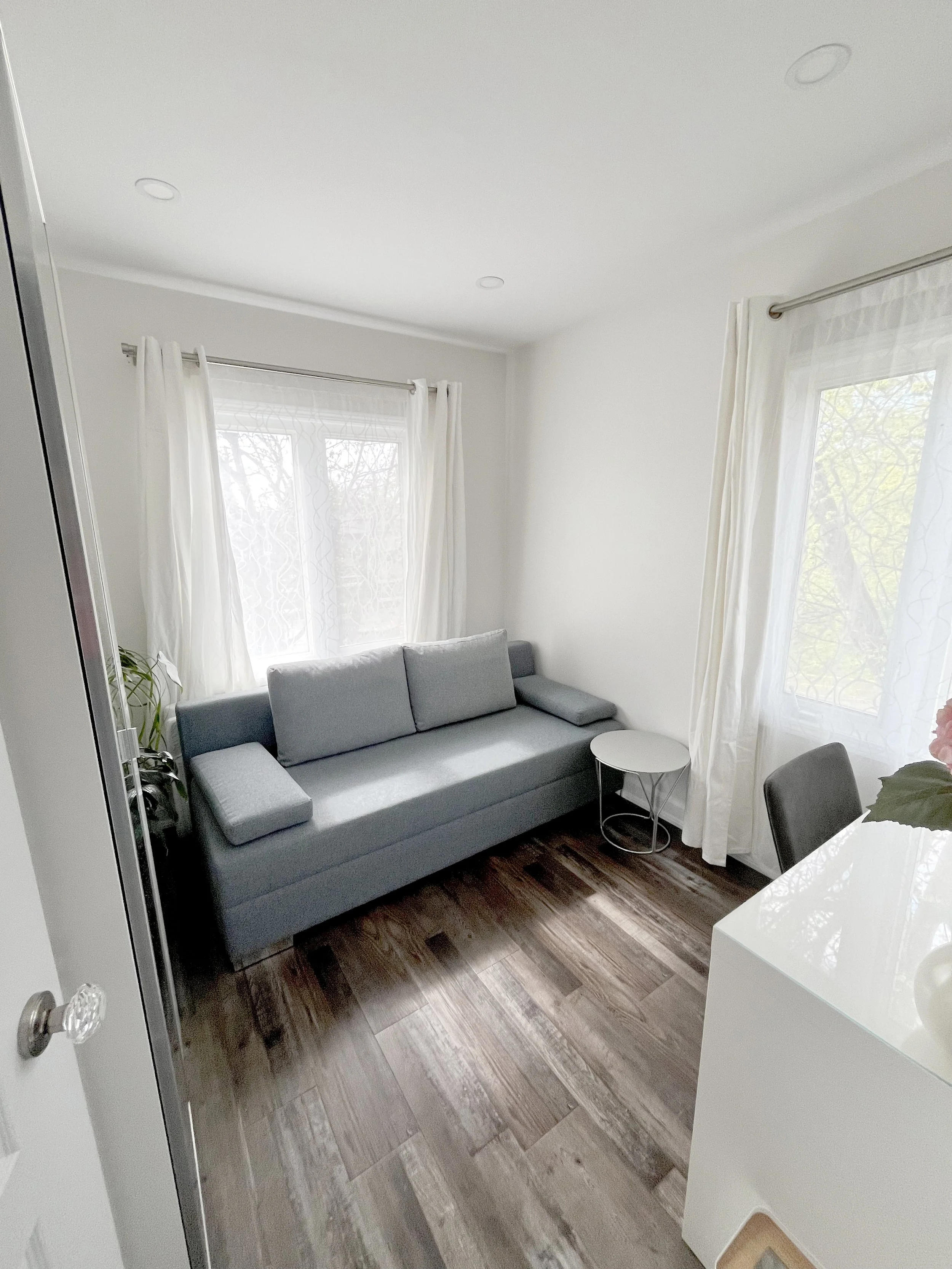 THE OASIS office and sitting room renovation with a grey sofa, sheer curtains, wood-look flooring, and a small round side table