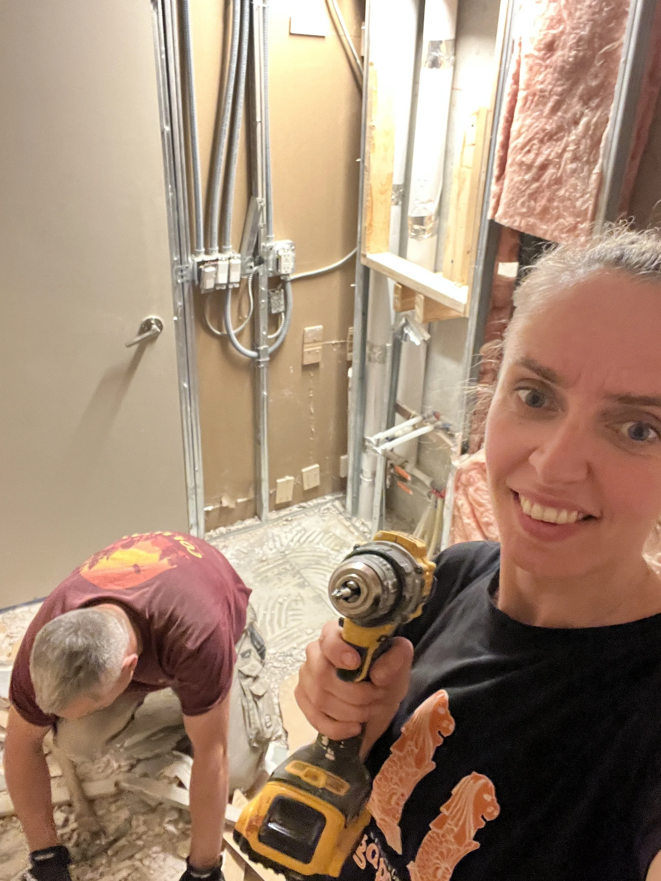 Val taking a selfie with a cordless drill while a crew member clears demolition debris behind her, with the bathroom wall stripped to steel studs and exposed electrical and plumbing, during the LUX NOIR bathroom renovation by Vogman Design + Build.