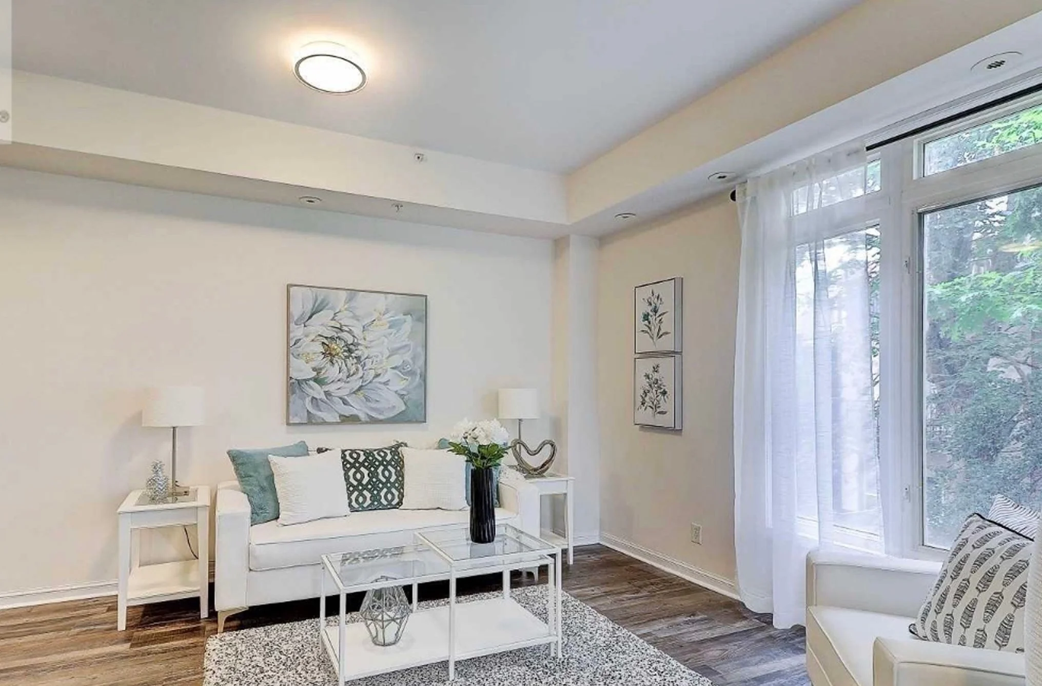 Before photo of THE OASIS living room, staged with a white sofa, floral wall art, and a glass coffee table, ahead of the full renovation