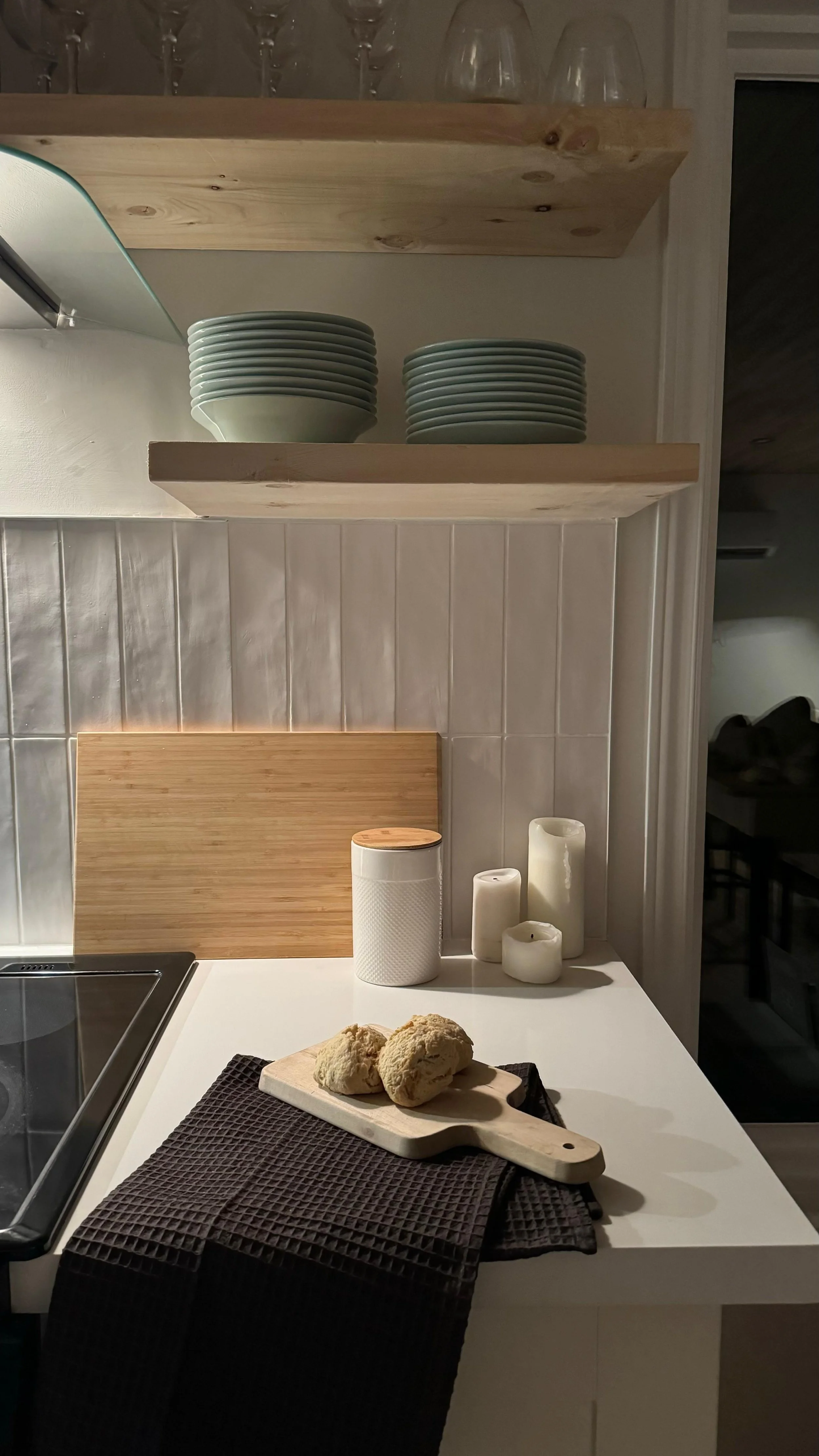 Styled kitchen corner in the Tobermory cottage renovation with open wood shelving, stacked sage green dishware, fluted white tile backsplash, and fresh bread on a cutting board