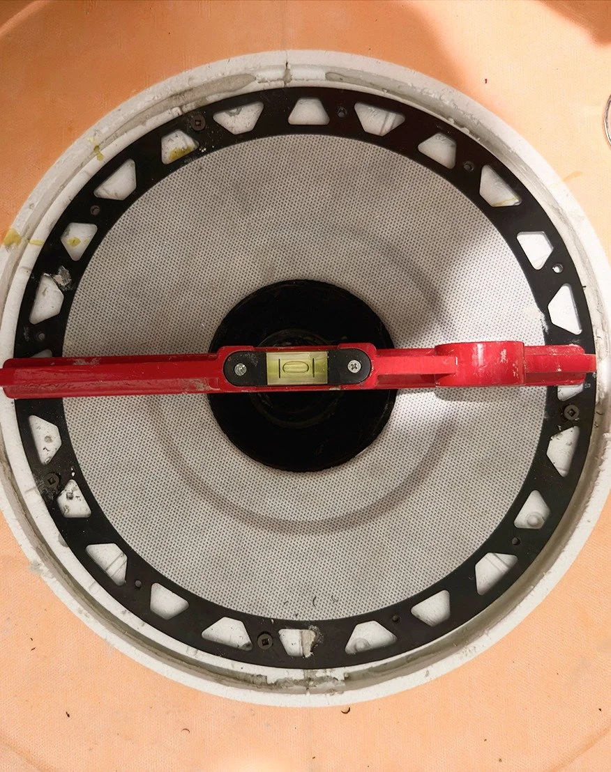 Overhead view of a red spirit level resting across a shower drain flange set into orange waterproofing membrane, checking that the drain is level, during The Rebuild renovation by Vogman Design + Build.