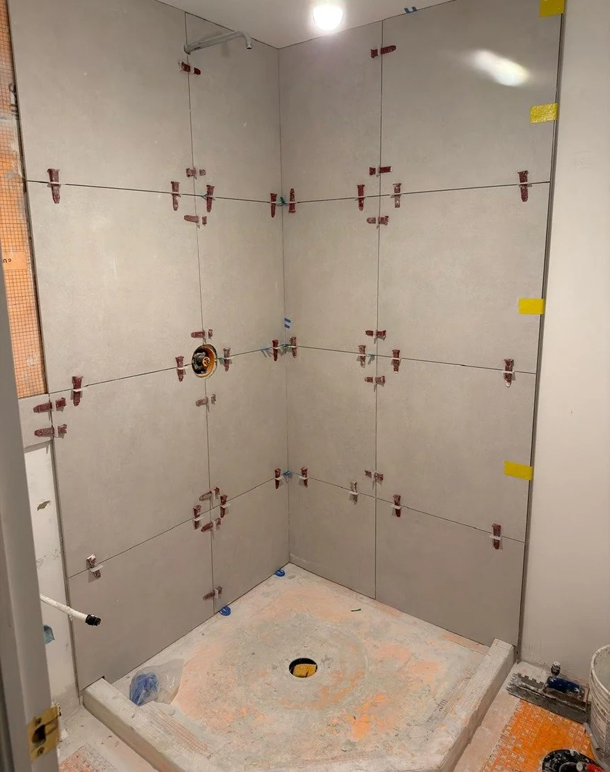 White bathroom vanity with an integrated rectangular sink and chrome single-handle faucet being installed against large-format light grey wall tile, with the drawers not yet fitted, during The Rebuild renovation by Vogman Design + Build.