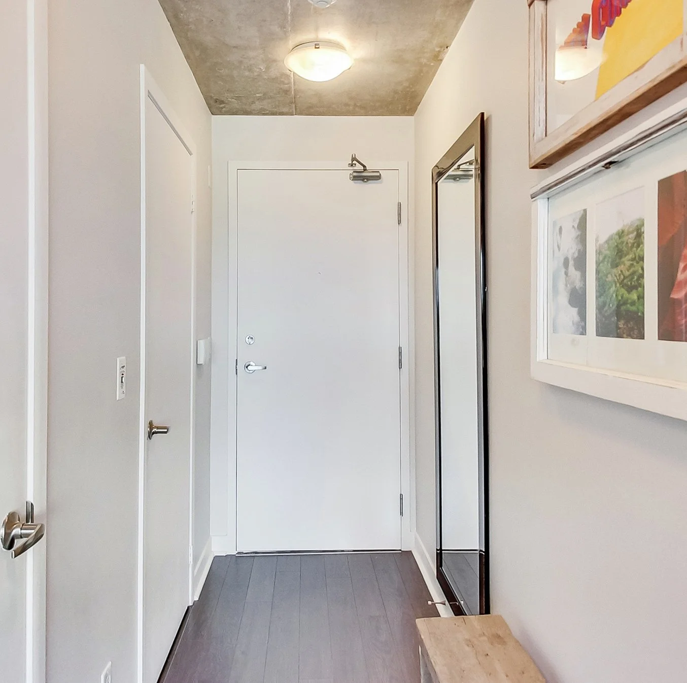 After photo of the iLOFT entryway with an exposed concrete ceiling, a full-length mirror, framed art, and dark wood flooring