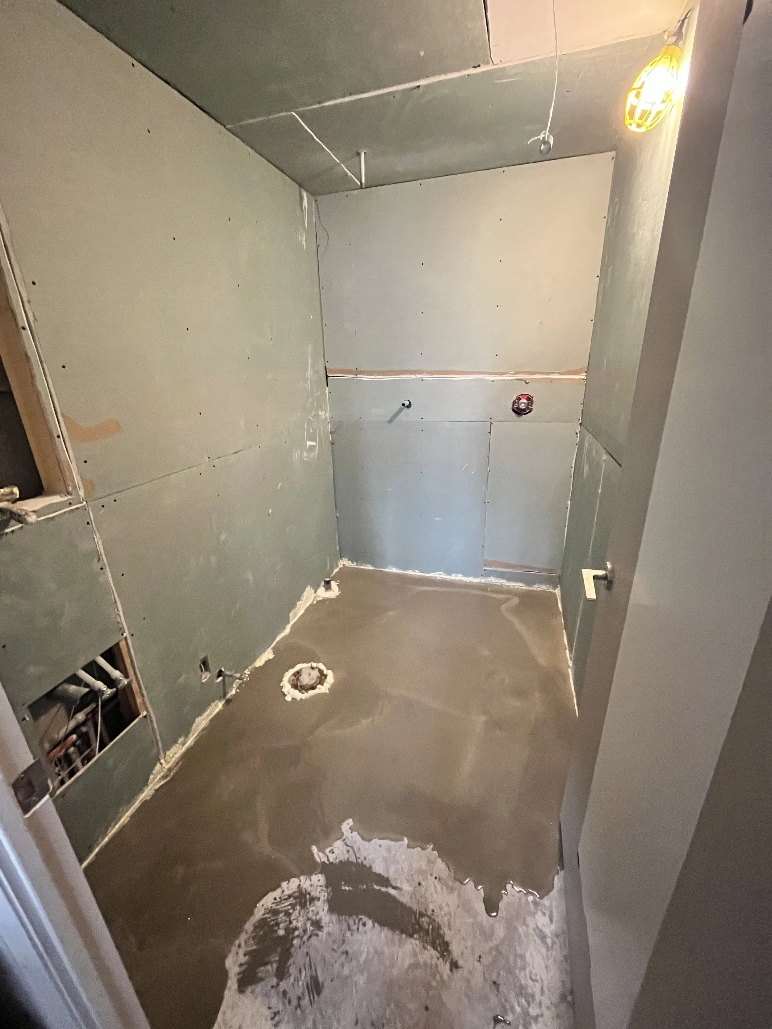 Freshly poured self-levelling compound spreading across the bathroom floor around the toilet flange, with moisture-resistant drywall on the walls and the shower valve roughed in, during the LUX NOIR bathroom renovation by Vogman Design + Build.