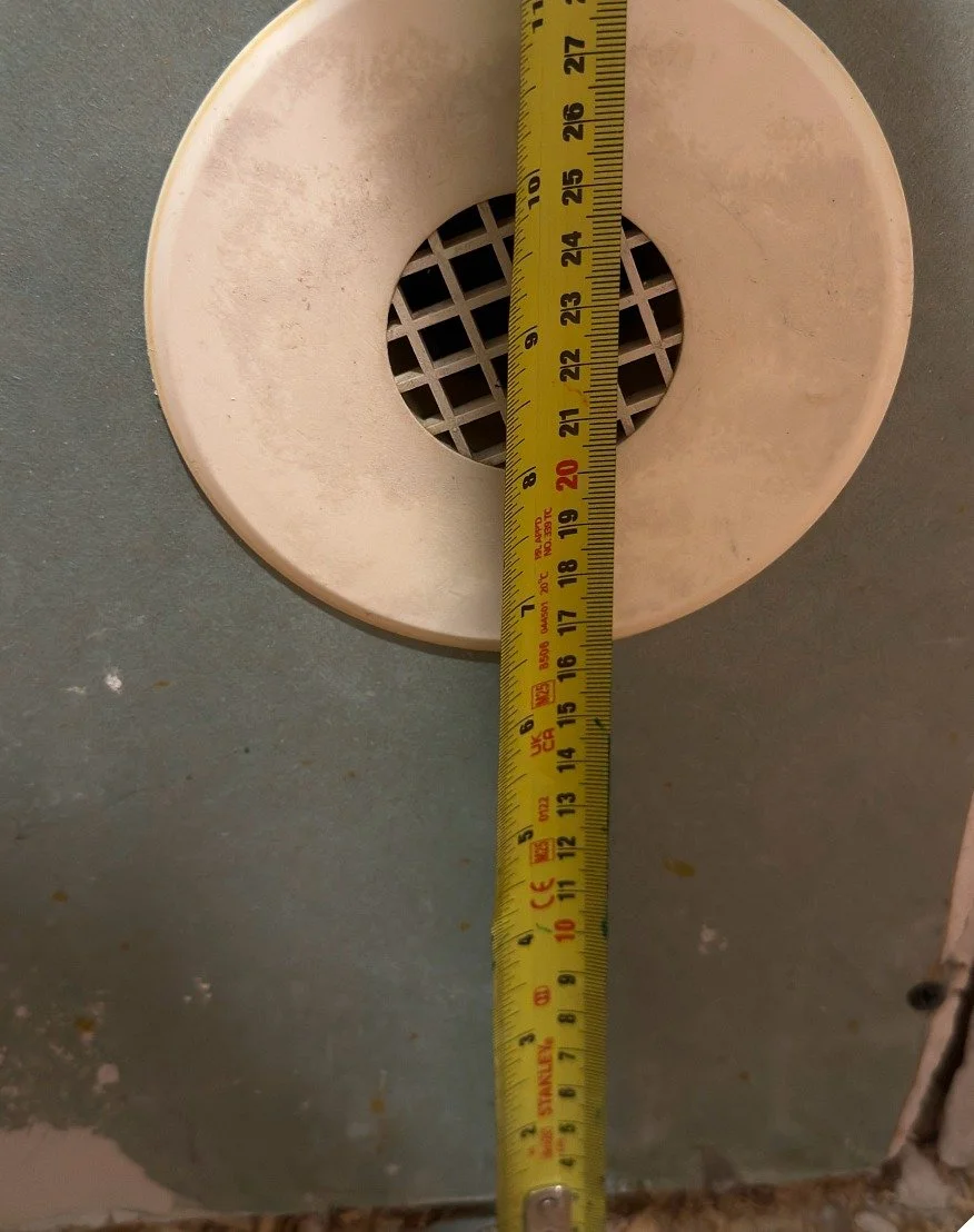 Round white floor air vent with a grille being measured with a yellow tape measure before being rerouted to the wall during The Rebuild renovation by Vogman Design + Build