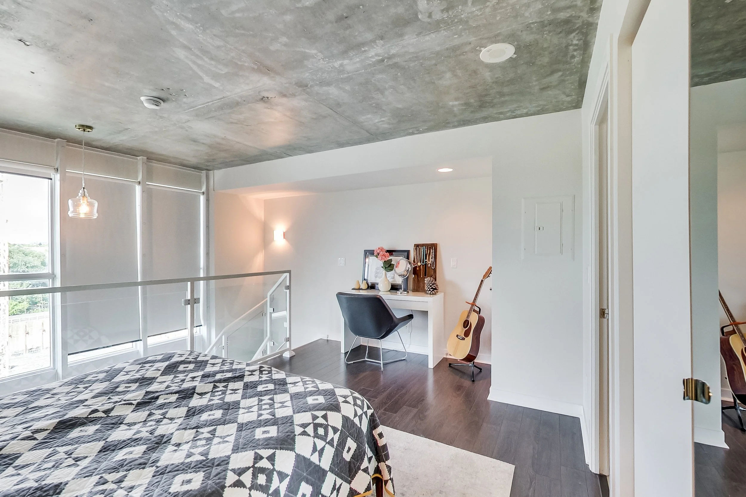 After photo of the iLOFT loft bedroom nook with a white vanity desk, black accent chair, guitar, and exposed concrete ceiling