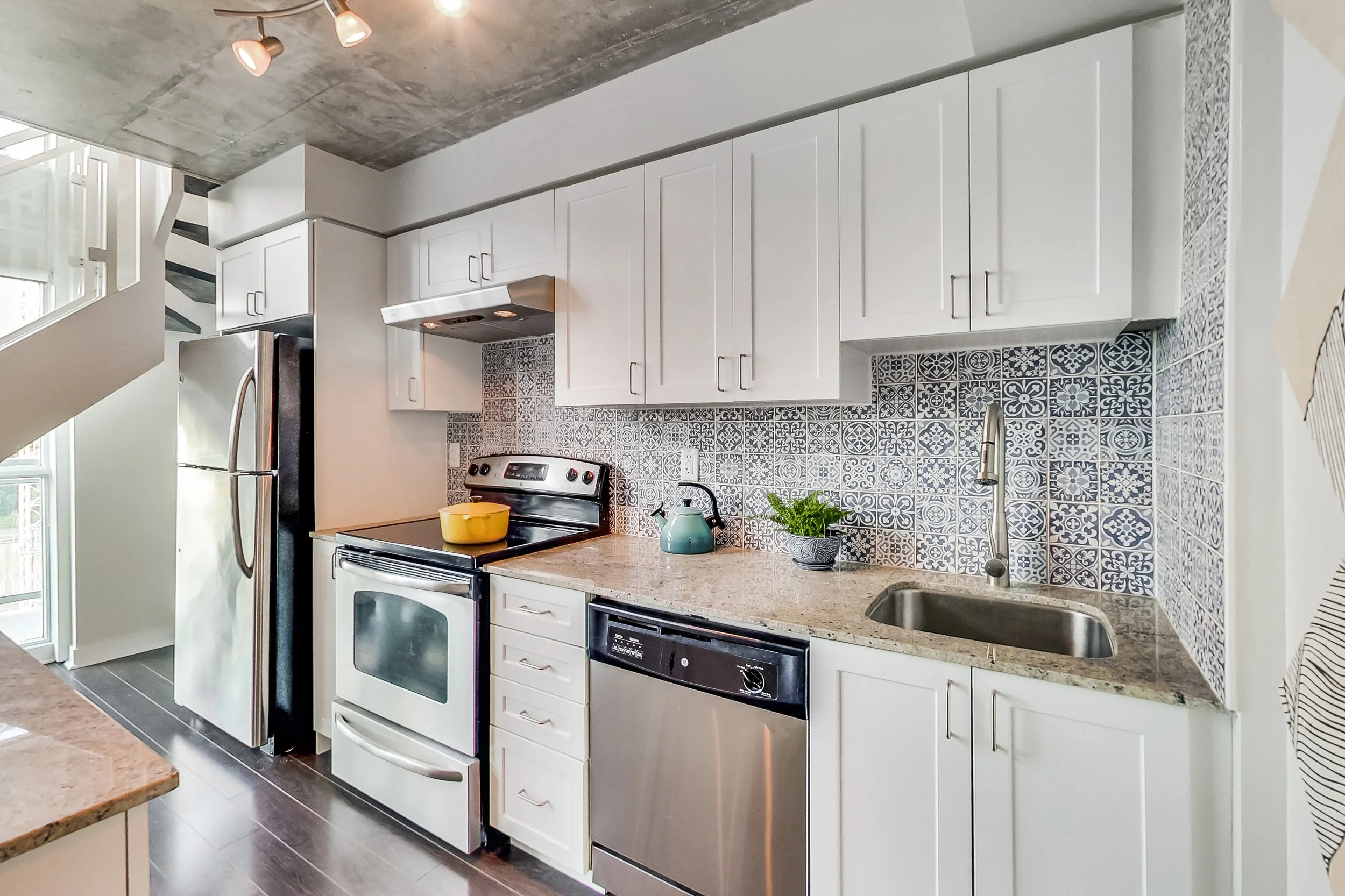 Renovated loft kitchen in Toronto with exposed concrete ceiling, white shaker cabinets, patterned blue and white Moroccan-tile backsplash, granite countertops, and stainless steel appliances