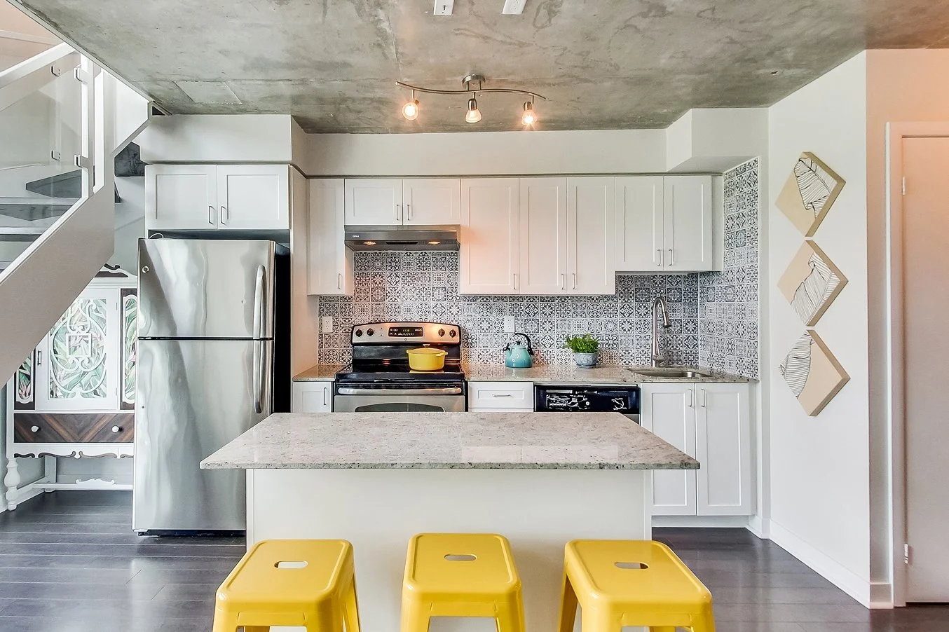 After photo of the iLOFT kitchen renovation with white cabinets, a patterned blue and white tile backsplash, exposed concrete ceiling, and yellow bar stools