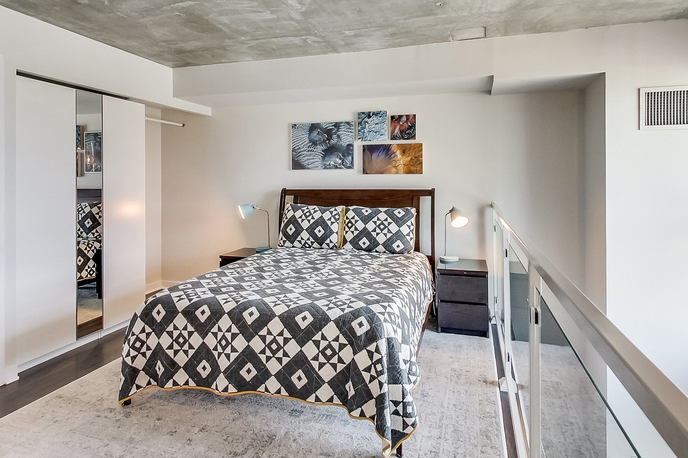 After photo of the iLOFT loft bedroom renovation with a dark wood bed frame, graphic diamond-patterned quilt, mirrored closet doors, and an exposed concrete ceiling