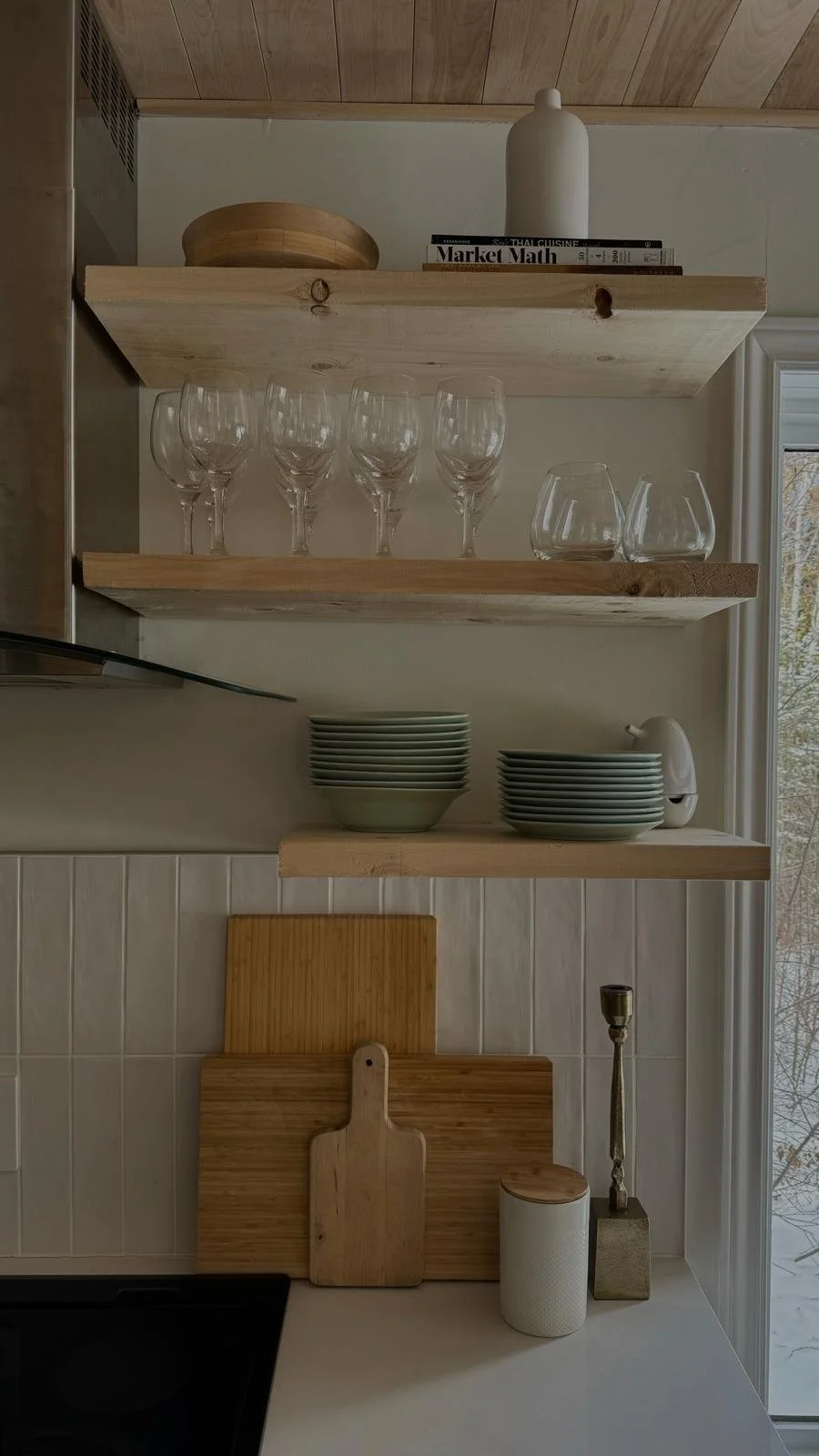 Open wood shelving in the Tobermory cottage kitchen styled with wine glasses, stacked ceramic bowls and plates, and wooden cutting boards against white tile
