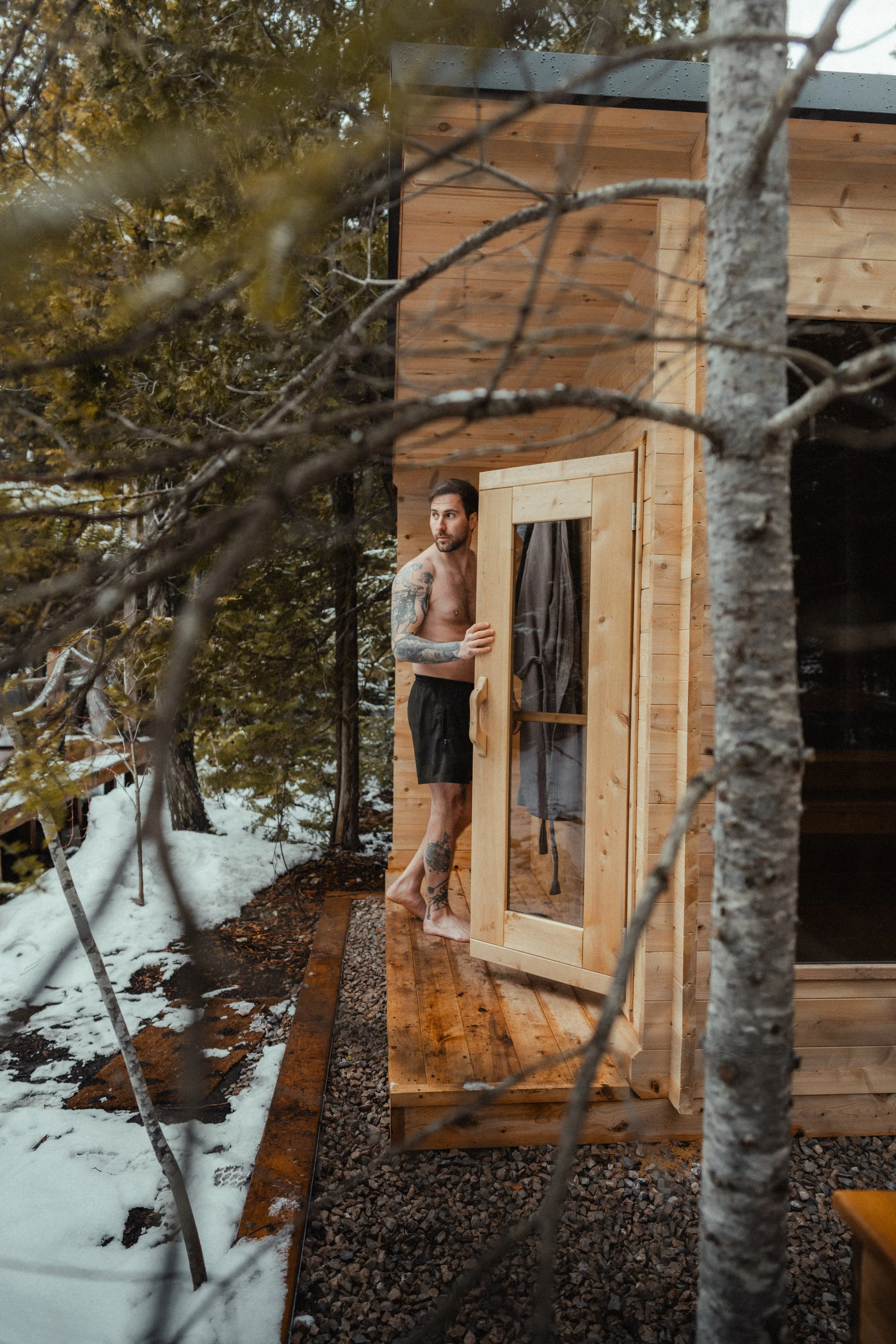 A person stepping out of a wood-clad outdoor sauna onto a snowy deck, surrounded by pine trees at the Tobermory cottage renovation