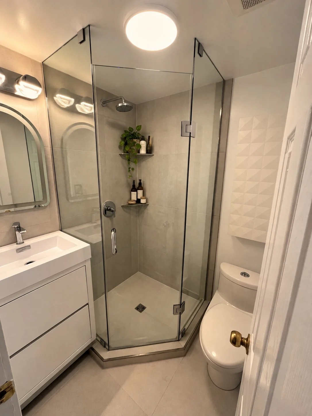 The Rebuild bathroom renovation with a corner frameless glass shower, built-in shower shelves, a white floating vanity, and a gold door handle