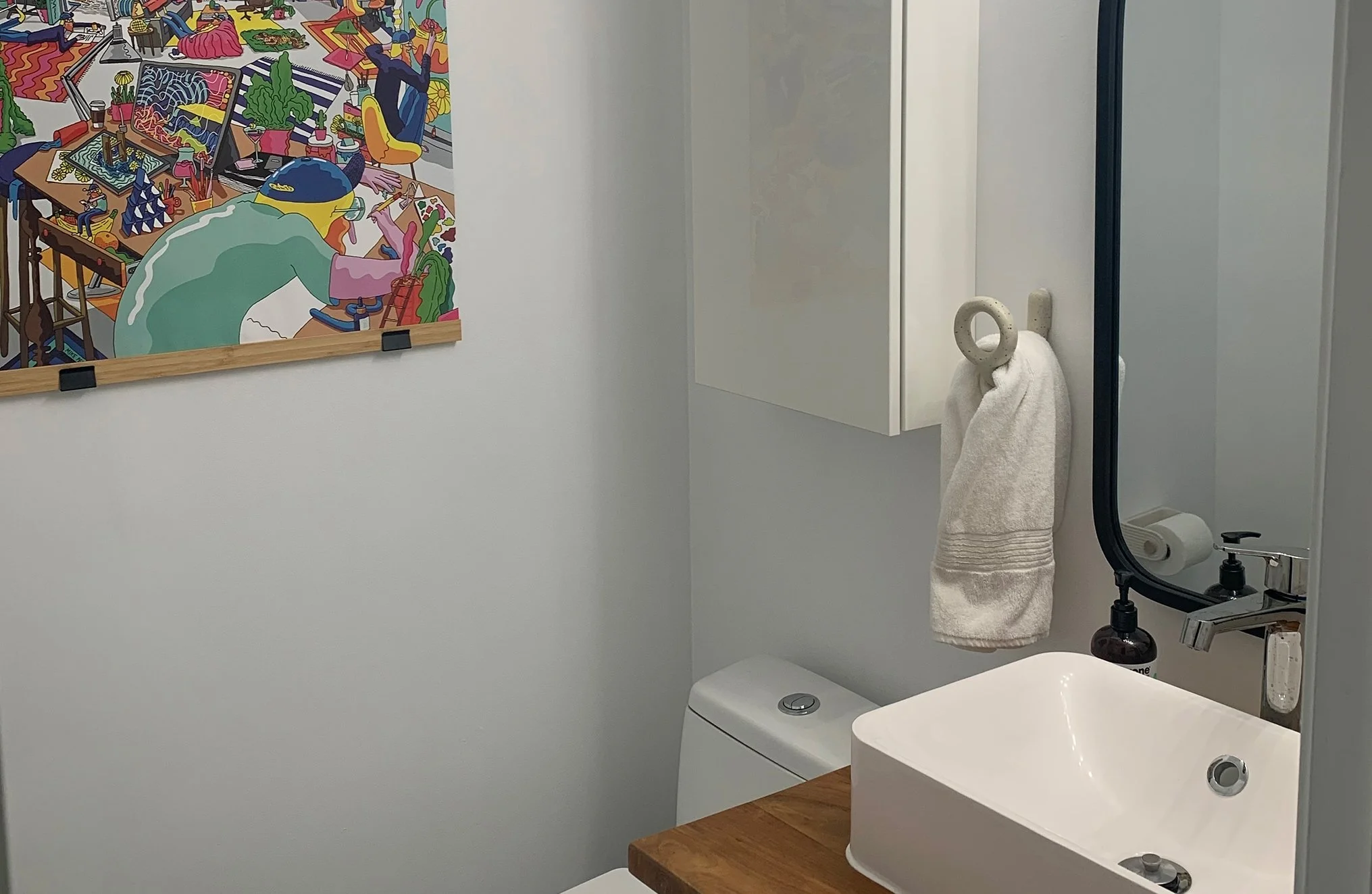 After photo of THE OASIS powder room renovation with a white vessel sink, wood countertop, black-framed mirror, and a colorful illustrated art print