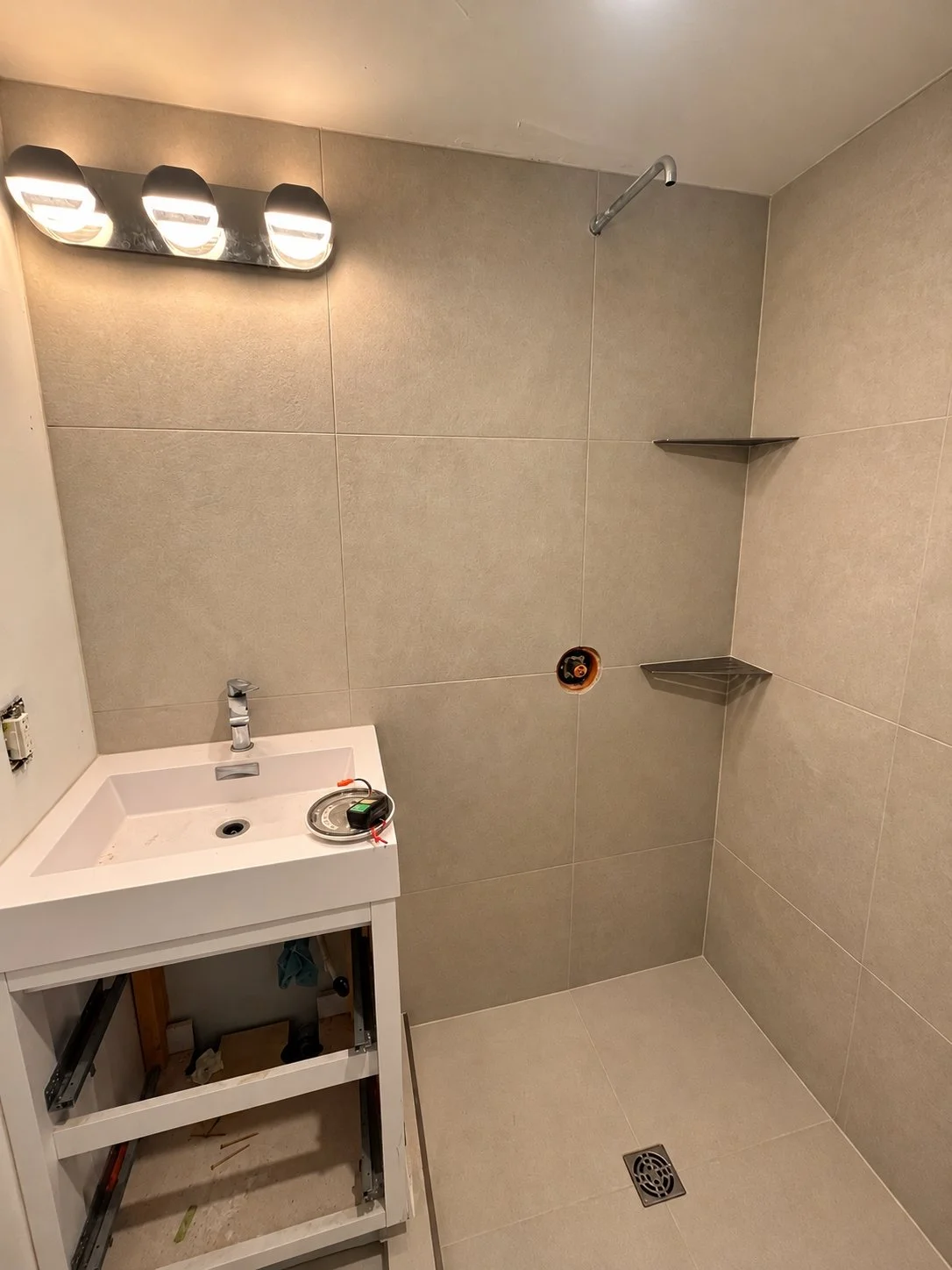 Bathroom mid-renovation with a white vanity being installed, tiled walk-in shower with built-in corner shelves, and a floor drain, during The Rebuild bathroom renovation