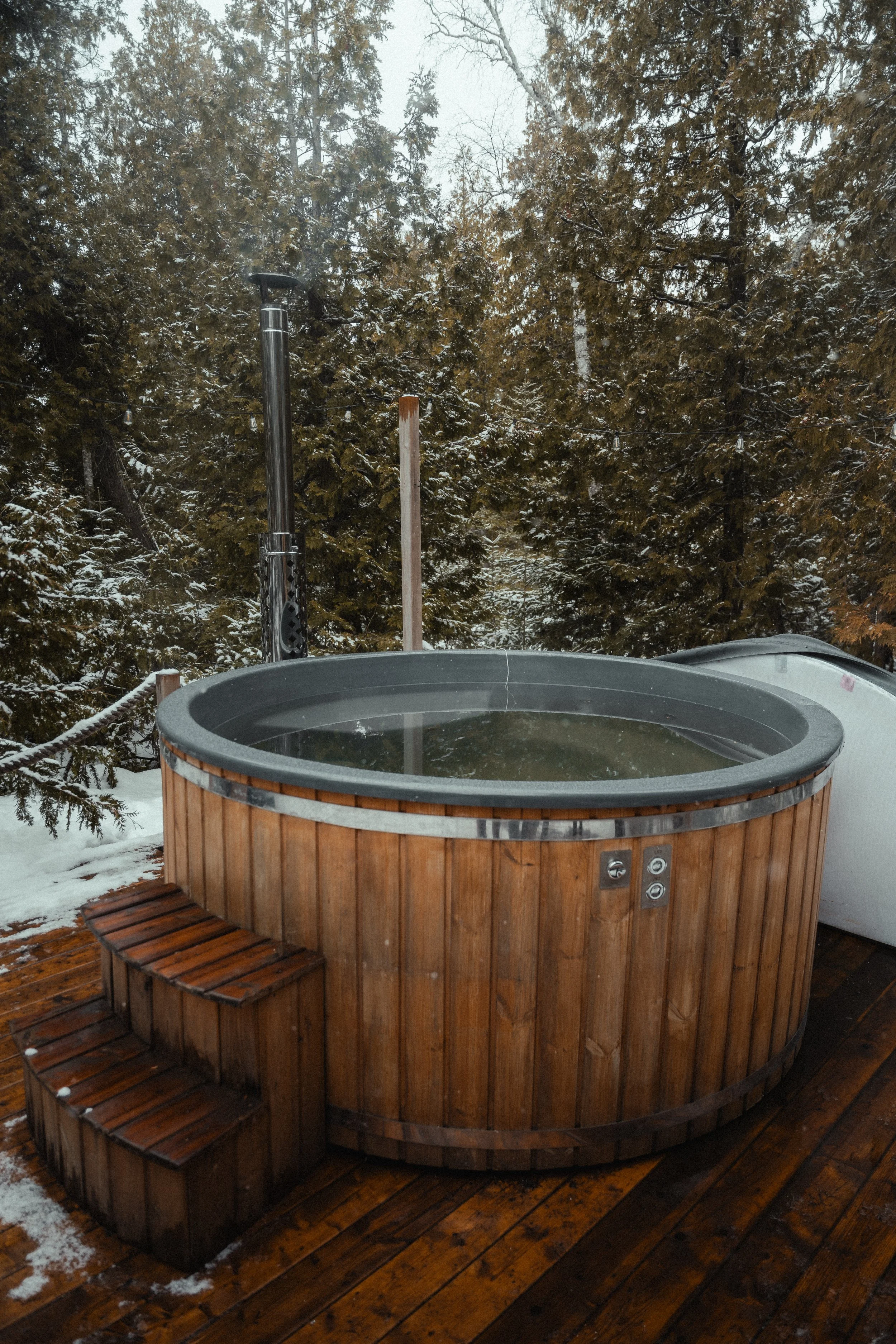 Close-up of a round wood-fired hot tub with a stainless steel chimney, steaming on a wood deck surrounded by snow-dusted pine trees