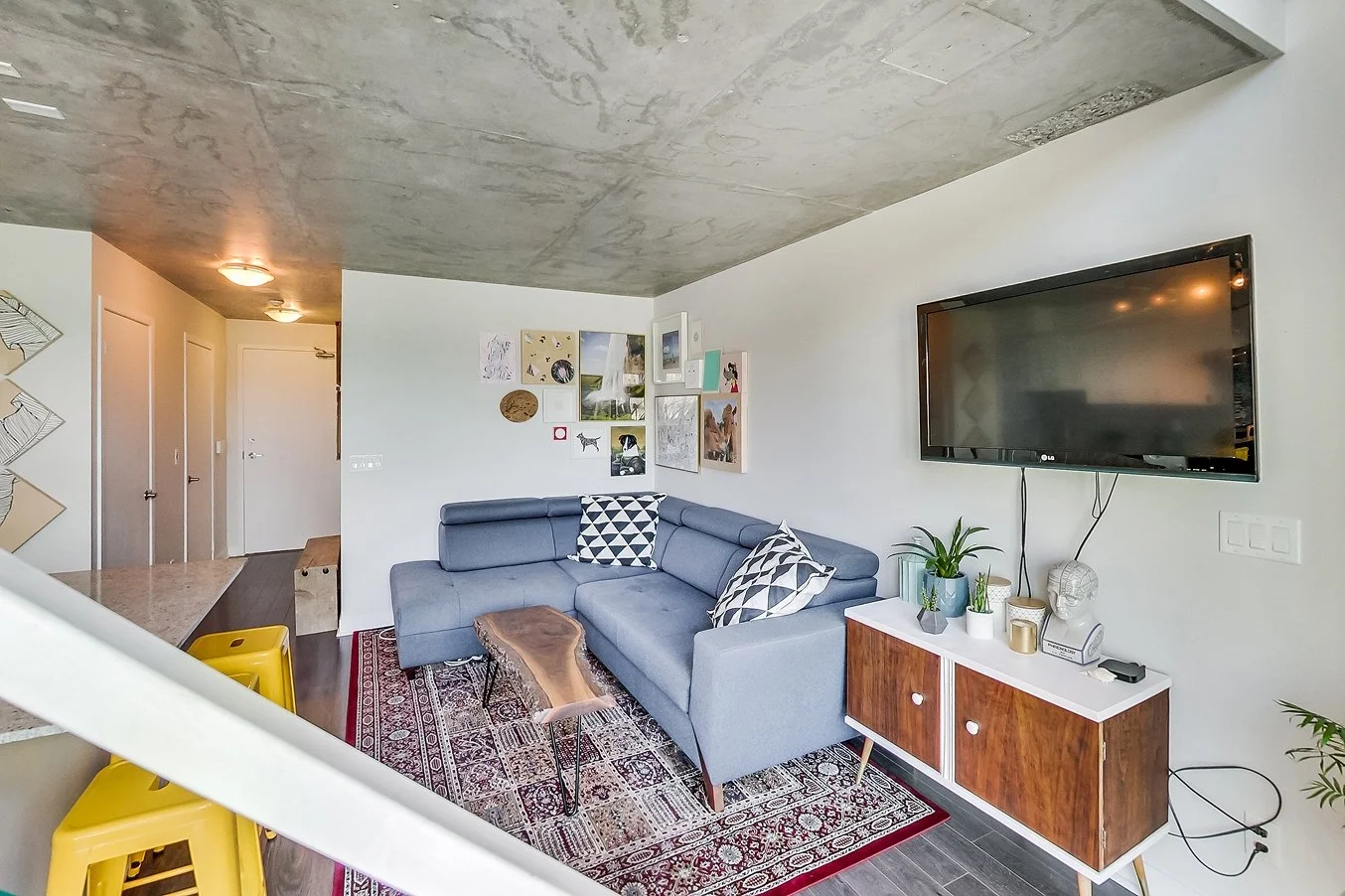 After photo of the iLOFT living room renovation with a blue sectional sofa, live-edge wood coffee table, patterned area rug, and a wall-mounted TV above a wood credenza
