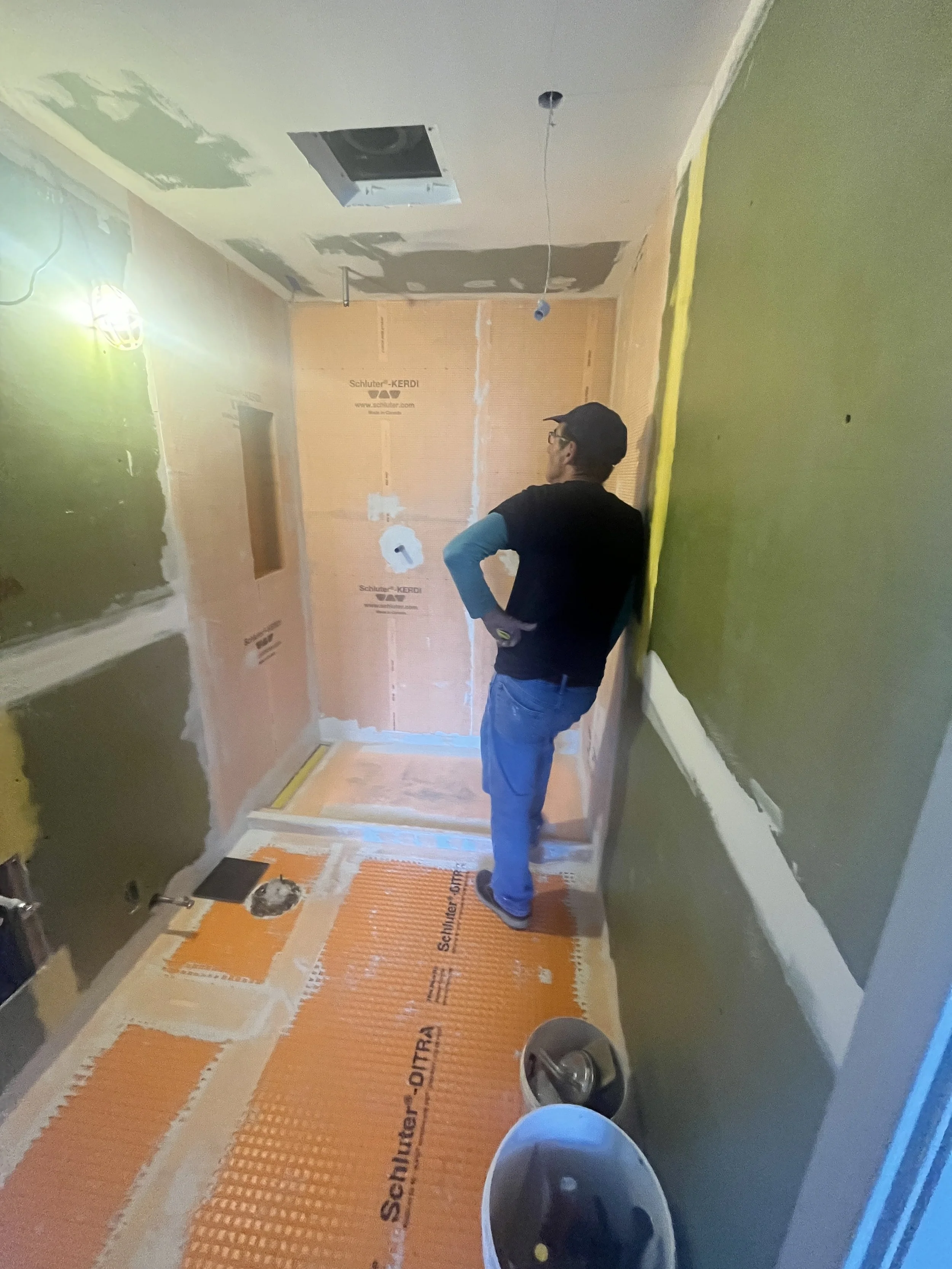 Petre surveying the bathroom before tiling, with orange Schluter-KERDI waterproofing membrane on the shower walls and niche and Schluter-DITRA uncoupling membrane on the floor, during the LUX NOIR bathroom renovation by Vogman Design + Build.