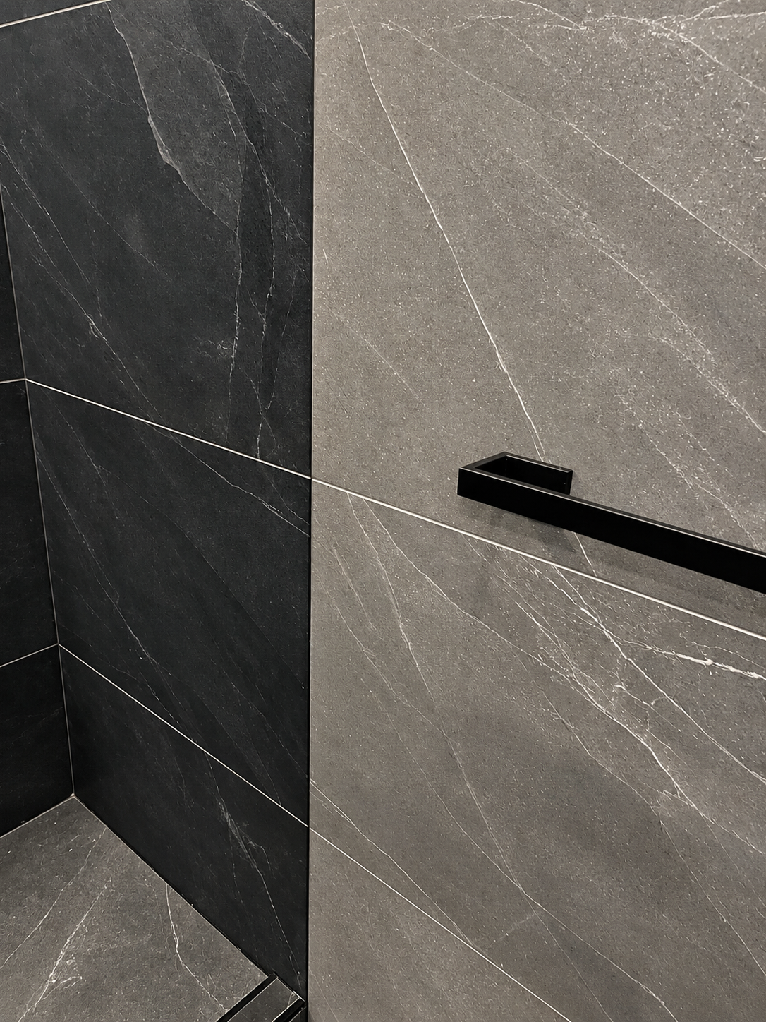 Close-up of charcoal stone-look shower tile meeting lighter grey veined wall tile at a clean vertical edge, with a matte black square towel bar, in the LUX NOIR bathroom renovation by Vogman Design + Build.