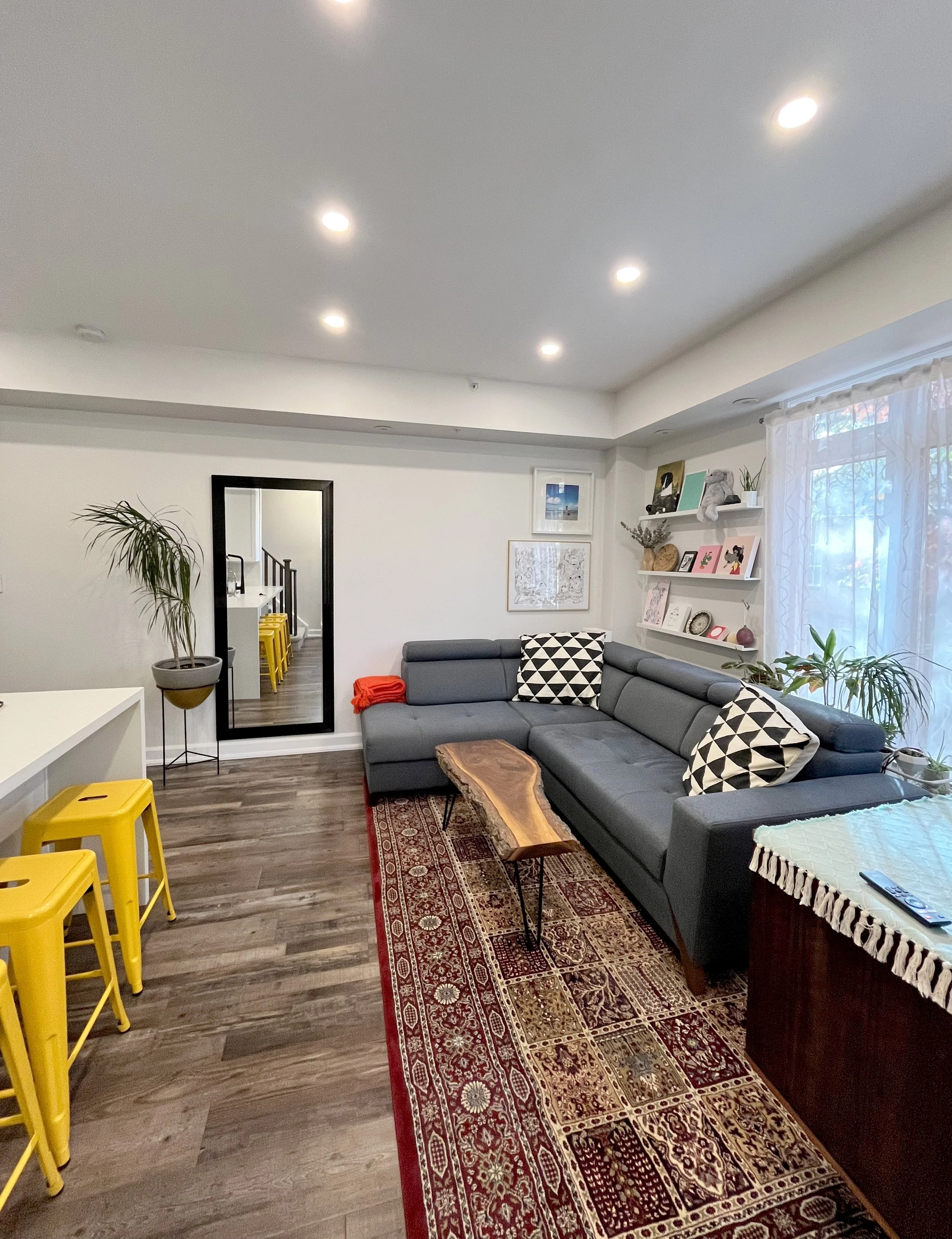 Eclectic living room with a grey leather sectional, red patterned area rug, live-edge wood coffee table, yellow bar stools, and open shelving styled with framed art and plants