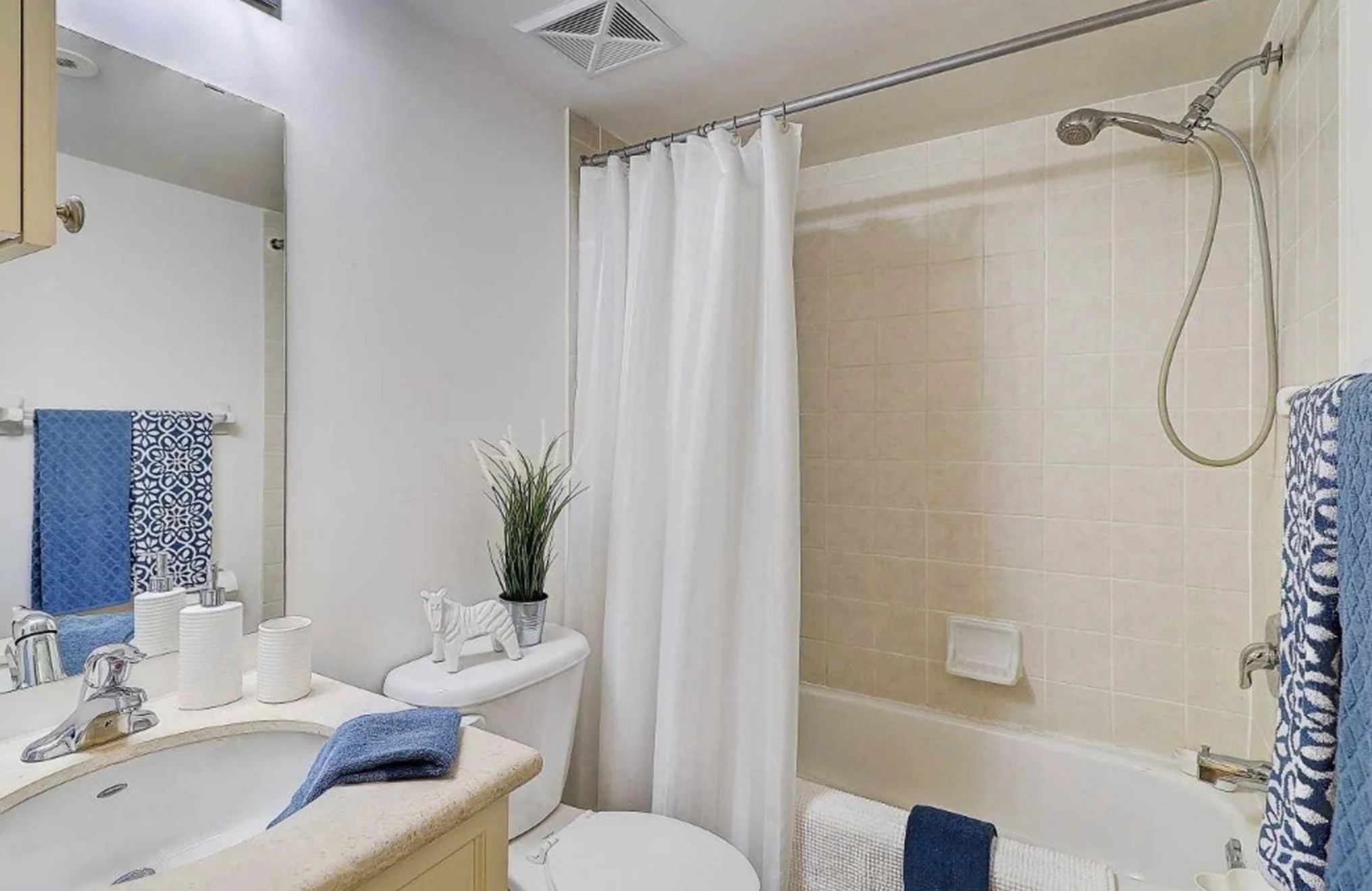 Before photo of THE OASIS bathroom with dated tan tile tub-shower combo, cream vanity, and blue patterned towels, ahead of the full renovation