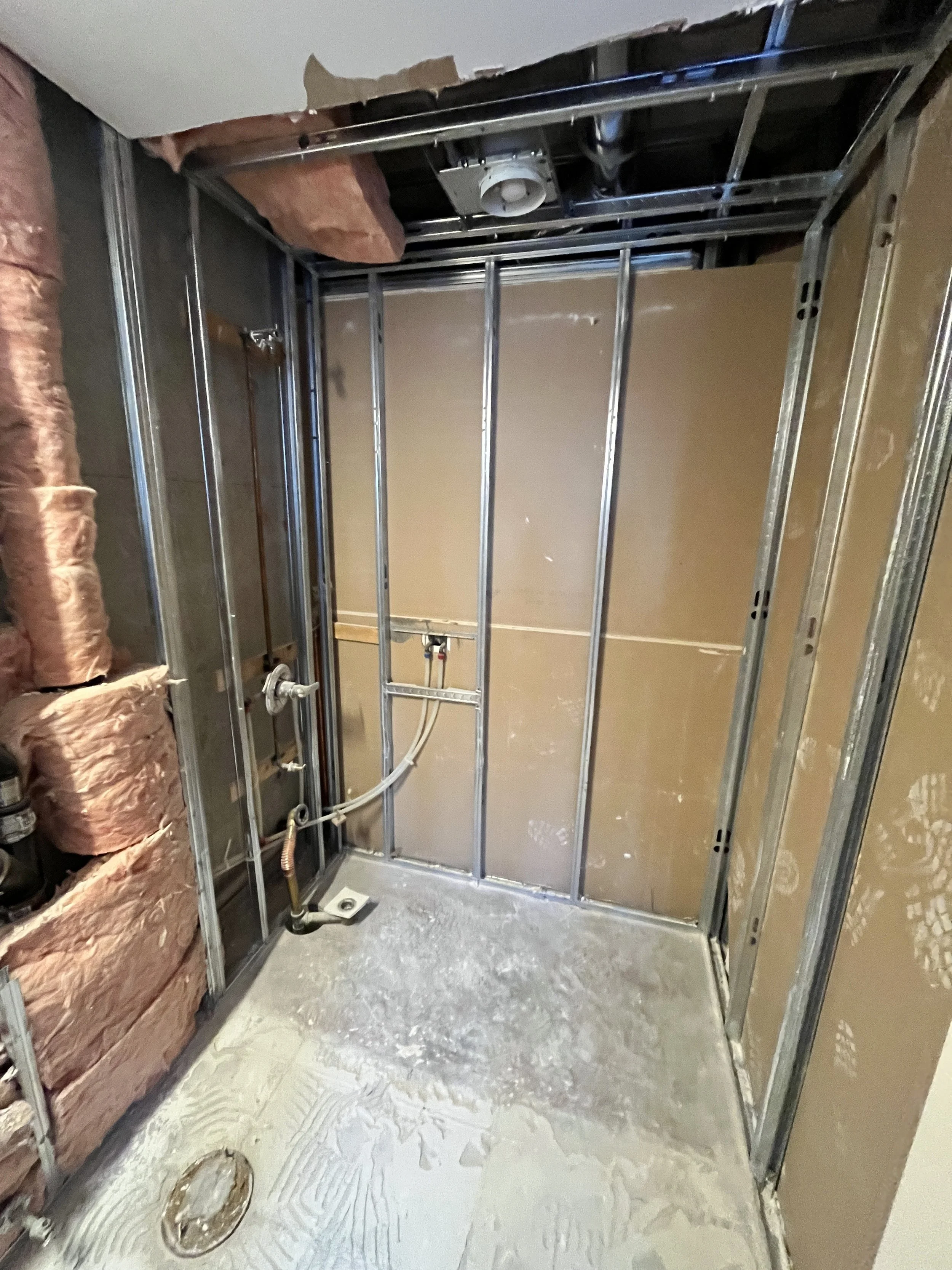 Bathroom fully demolished down to the steel studs and concrete floor, with the original plumbing lines, tub drain, toilet flange, insulation and ceiling exhaust fan exposed, during the LUX NOIR bathroom renovation by Vogman Design + Build.