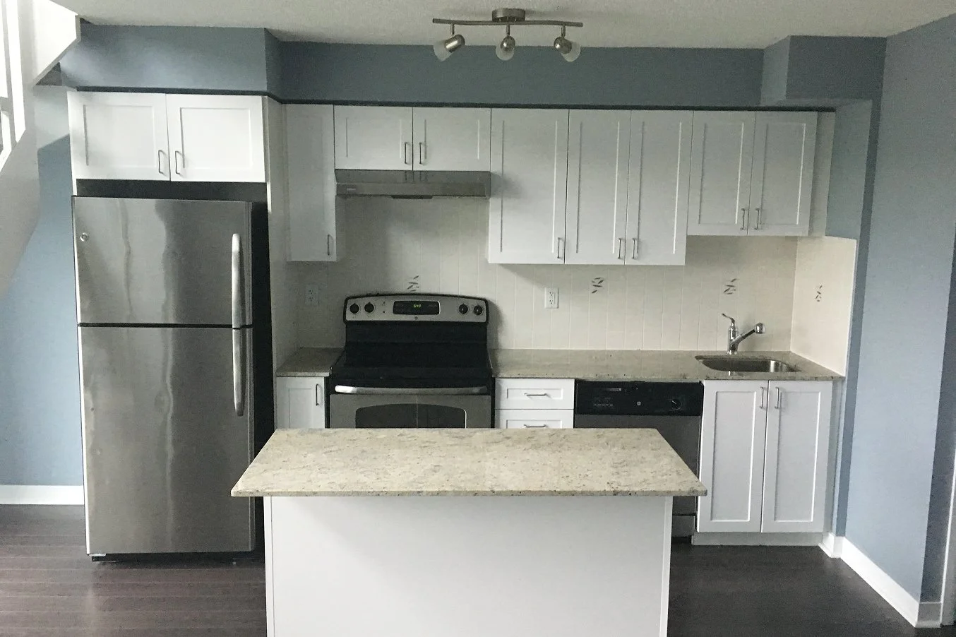 Before photo of a kitchen with blue walls, white cabinets, stainless steel appliances, and a granite countertop island, ahead of renovation