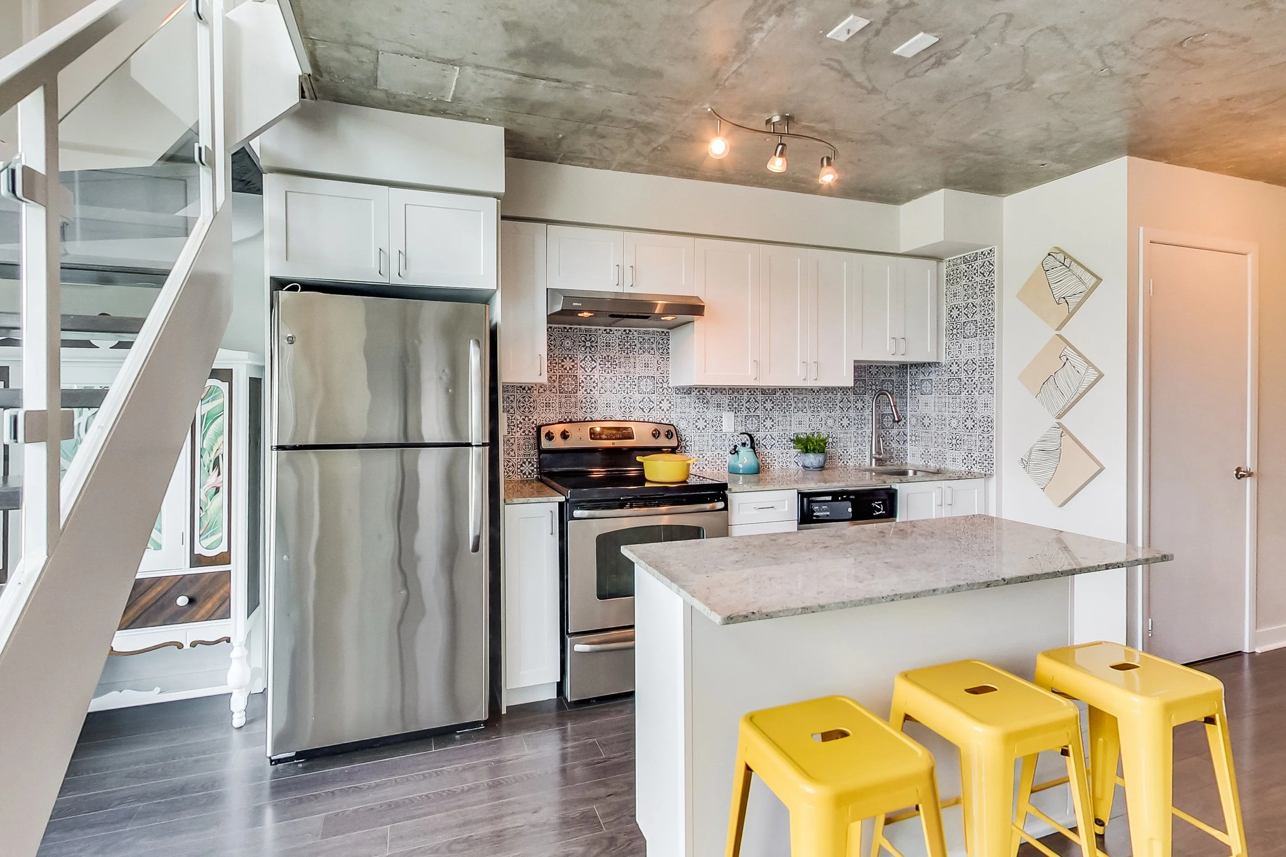 iLOFT kitchen in Etobicoke with white cabinets, a patterned blue and white tile backsplash, exposed concrete ceiling, stainless steel appliances, and yellow bar stools