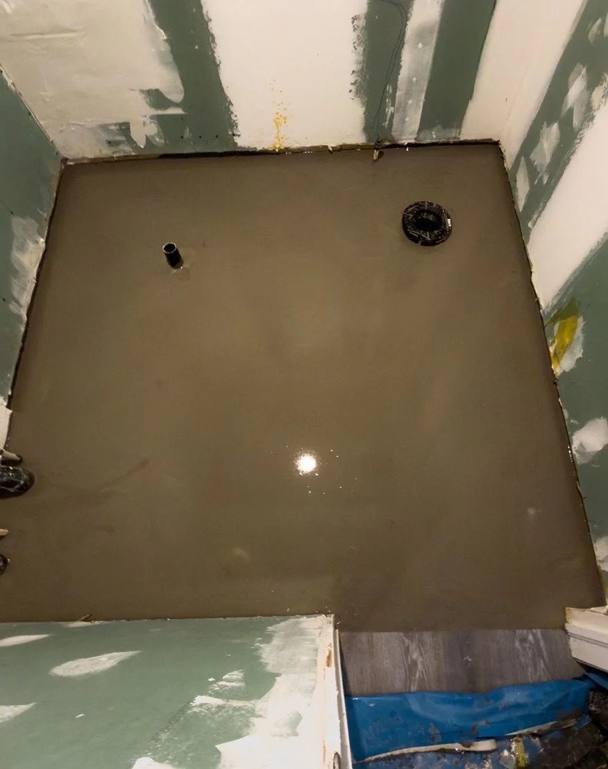Freshly poured self-levelling compound covering a bathroom floor, with the toilet flange and a drain pipe exposed and taped drywall on the surrounding walls, during The Rebuild renovation by Vogman Design + Build
