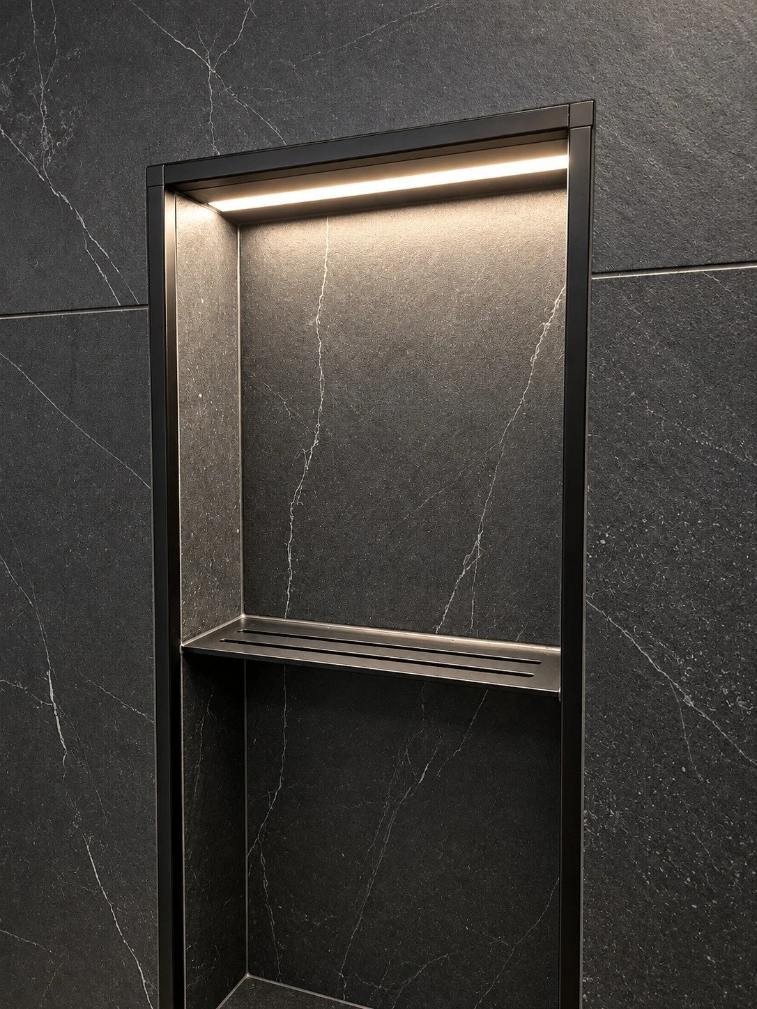 Recessed shower niche framed in matte black metal trim with a slotted metal shelf and built-in LED lighting, set into charcoal stone-look tile with white veining, in the LUX NOIR bathroom renovation by Vogman Design + Build.