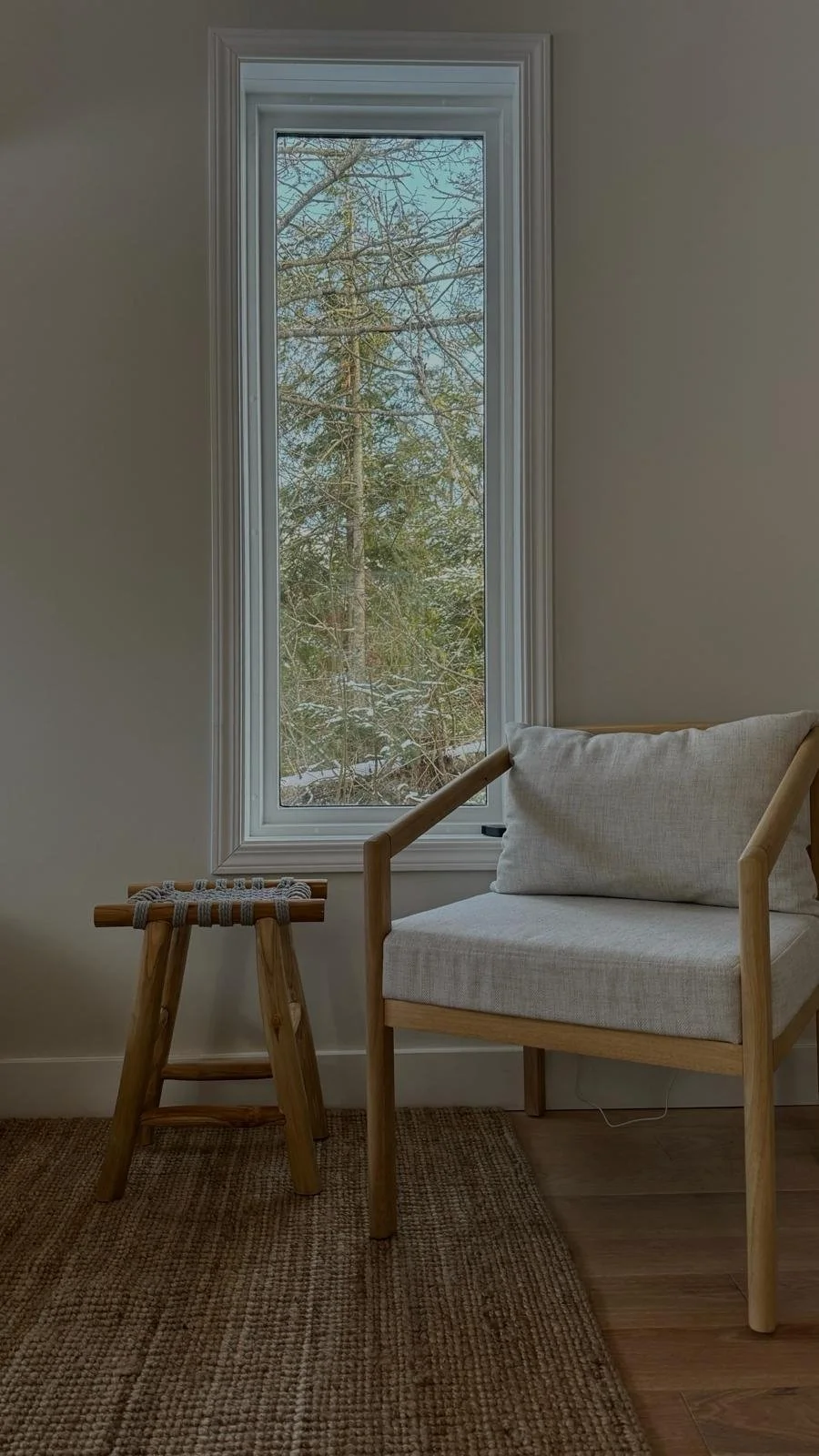 Cozy reading nook in the Tobermory cottage renovation with a tall wood-framed window overlooking snowy trees, a wood-framed accent chair, and a woven stool - Vogman Design + Build.