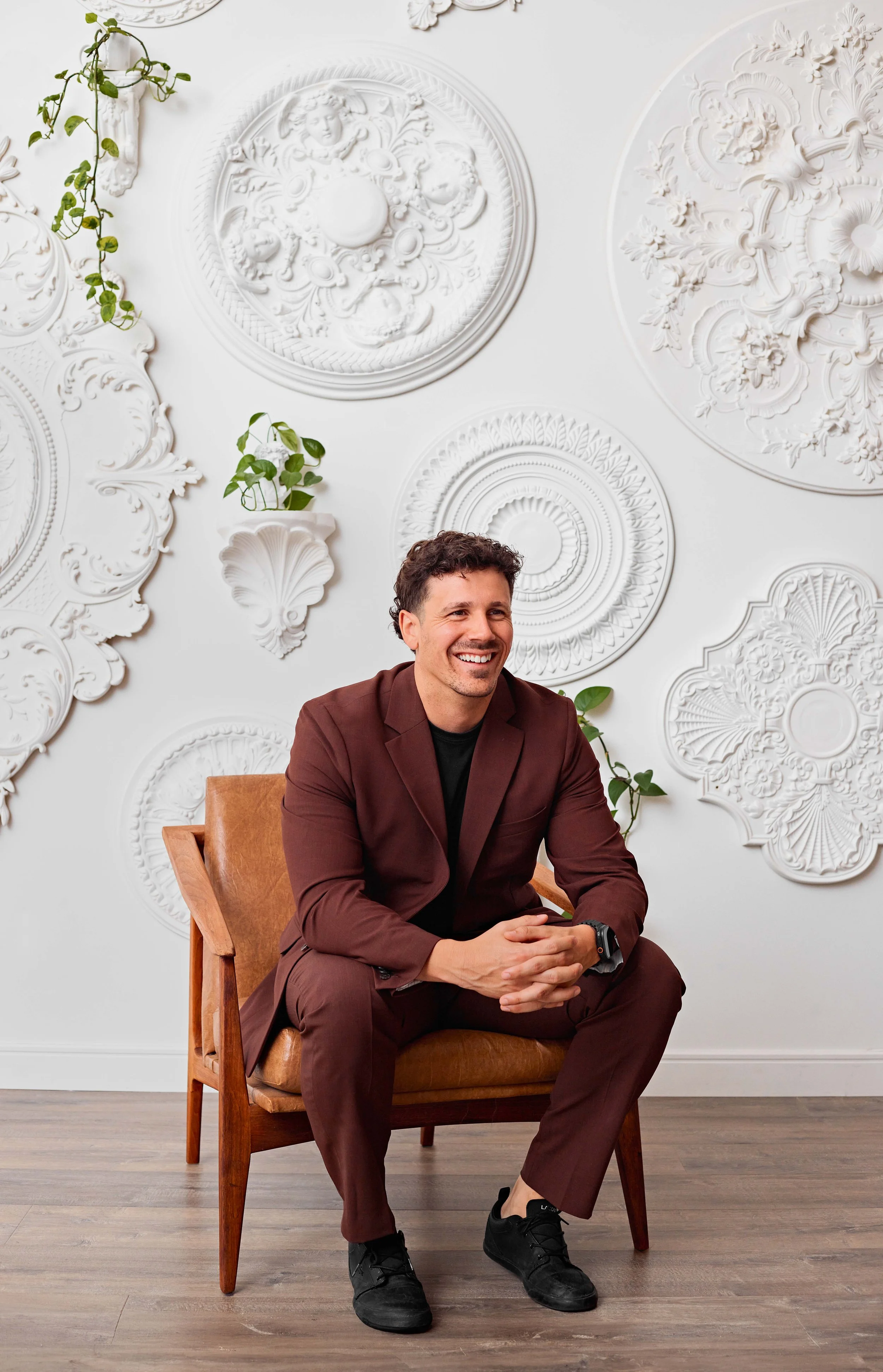 Olivier Bourgoin Courtier Immobilier souriant assis sur une chaise en bois, portant un costume marron, dans une pièce avec un mur blanc décoré de moulures et d'ornements en relief, avec des plantes vertes suspendues.