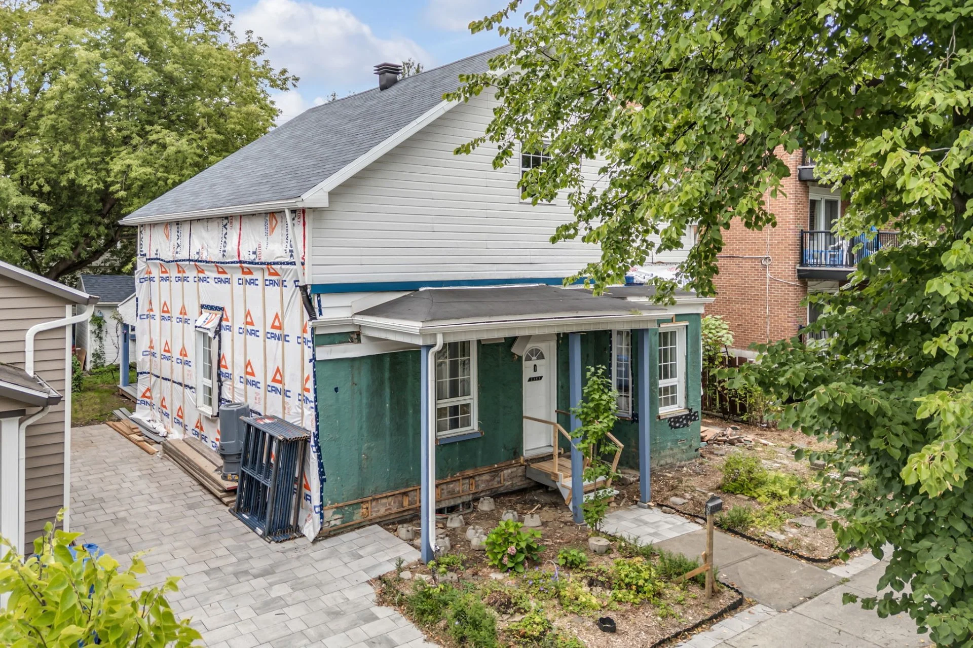 Une maison en cours de rénovation avec des travaux d'isolation en façade, entourée d'arbres et d'espaces verts, avec deux niveaux, un porche blanc, et une allée pavée devant.