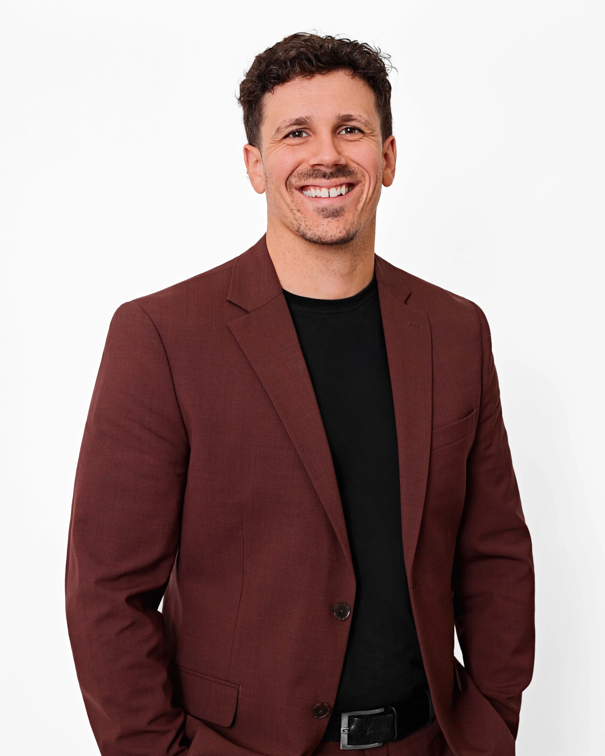 Olivier Bourgoin Courtier Immobilier souriant porte un costume marron foncé avec une chemise noire, debout contre un fond blanc.