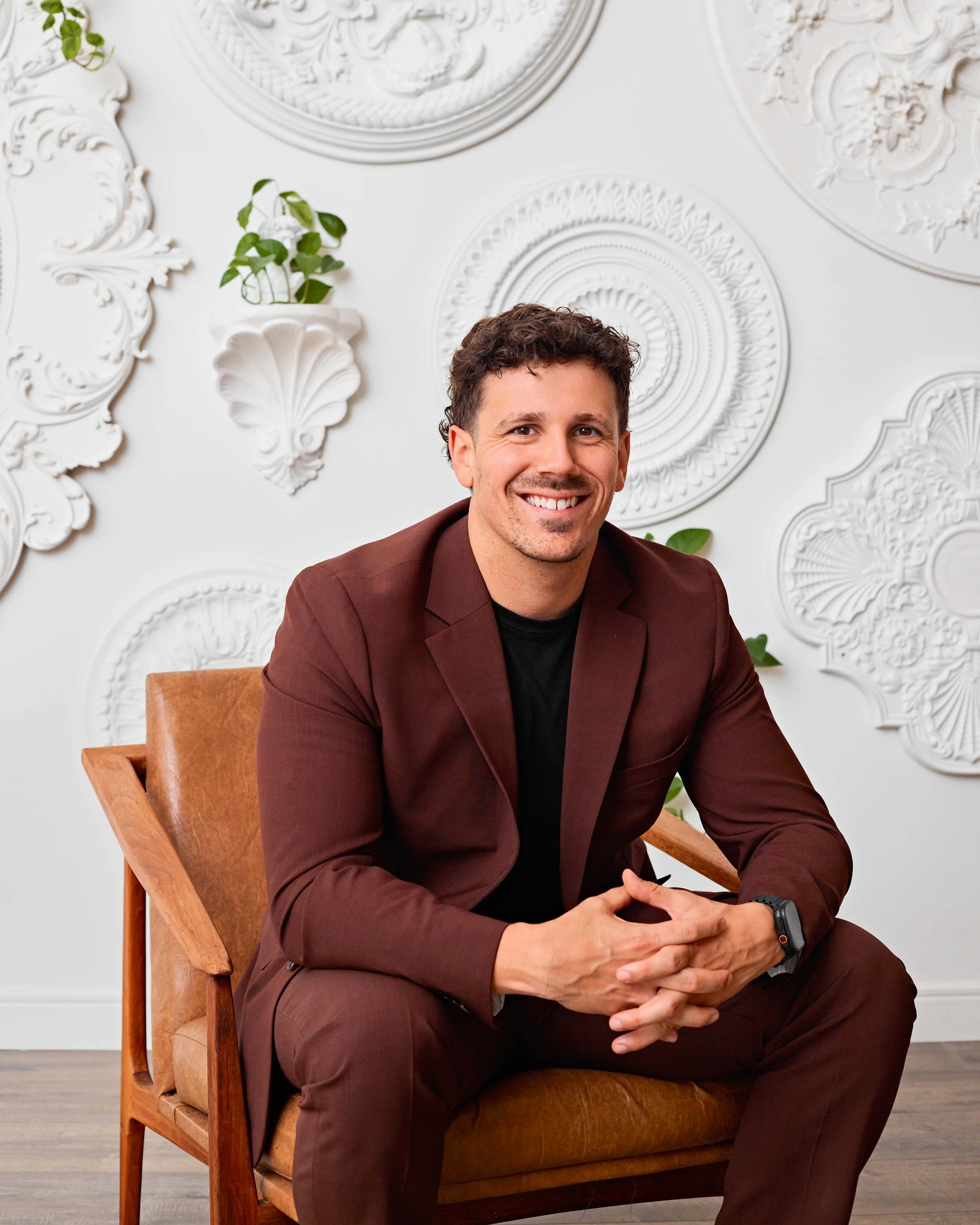 Olivier Bourgoin Courtier Immobilier souriant en costume marron, assis sur une chaise en bois dans une pièce avec un mur blanc orné de moulures décoratives et quelques plantes vertes.
