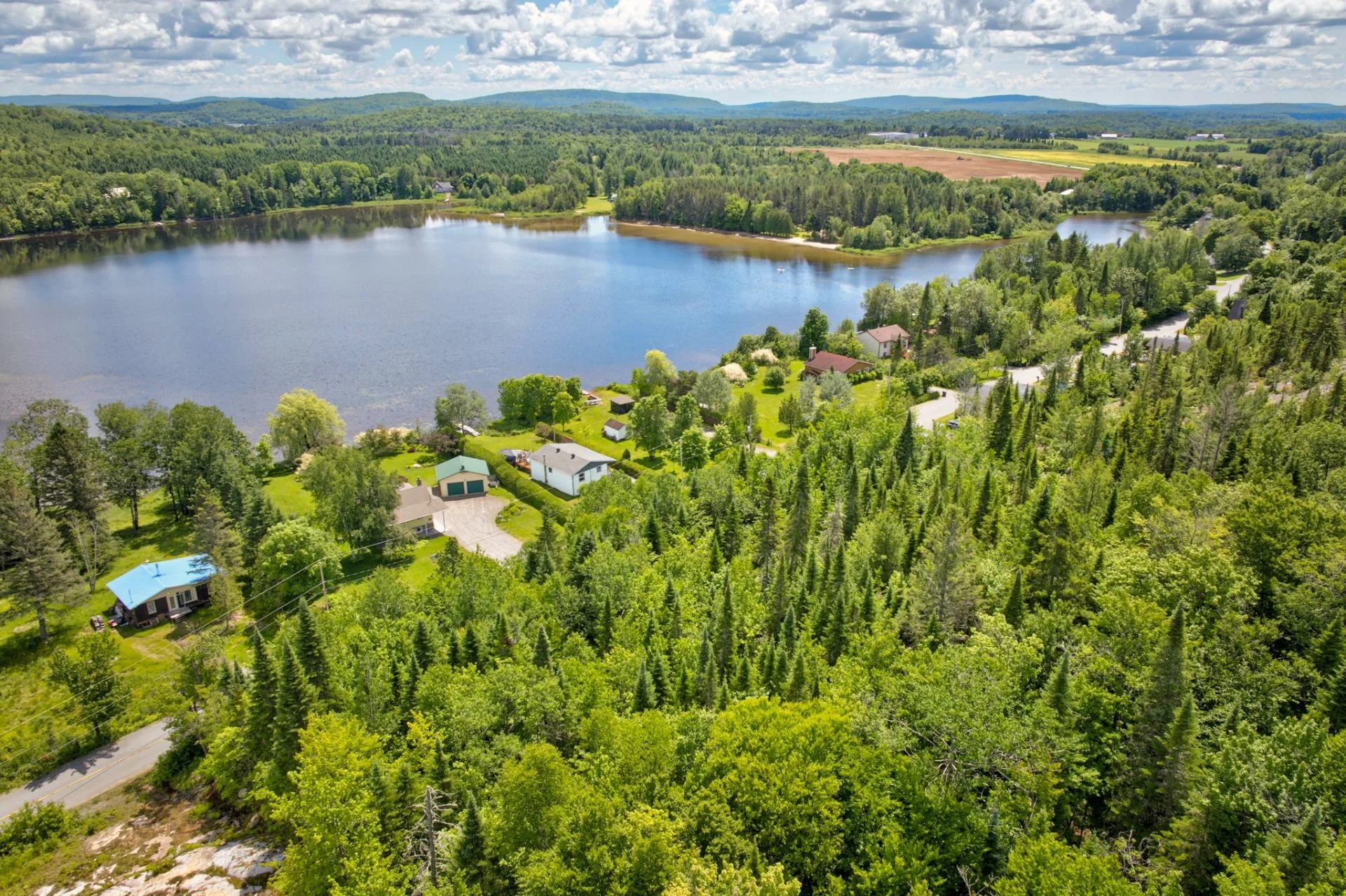 Vue aérienne d'un lac entouré de forêts et de maisons, avec un ciel partiellement nuageux.