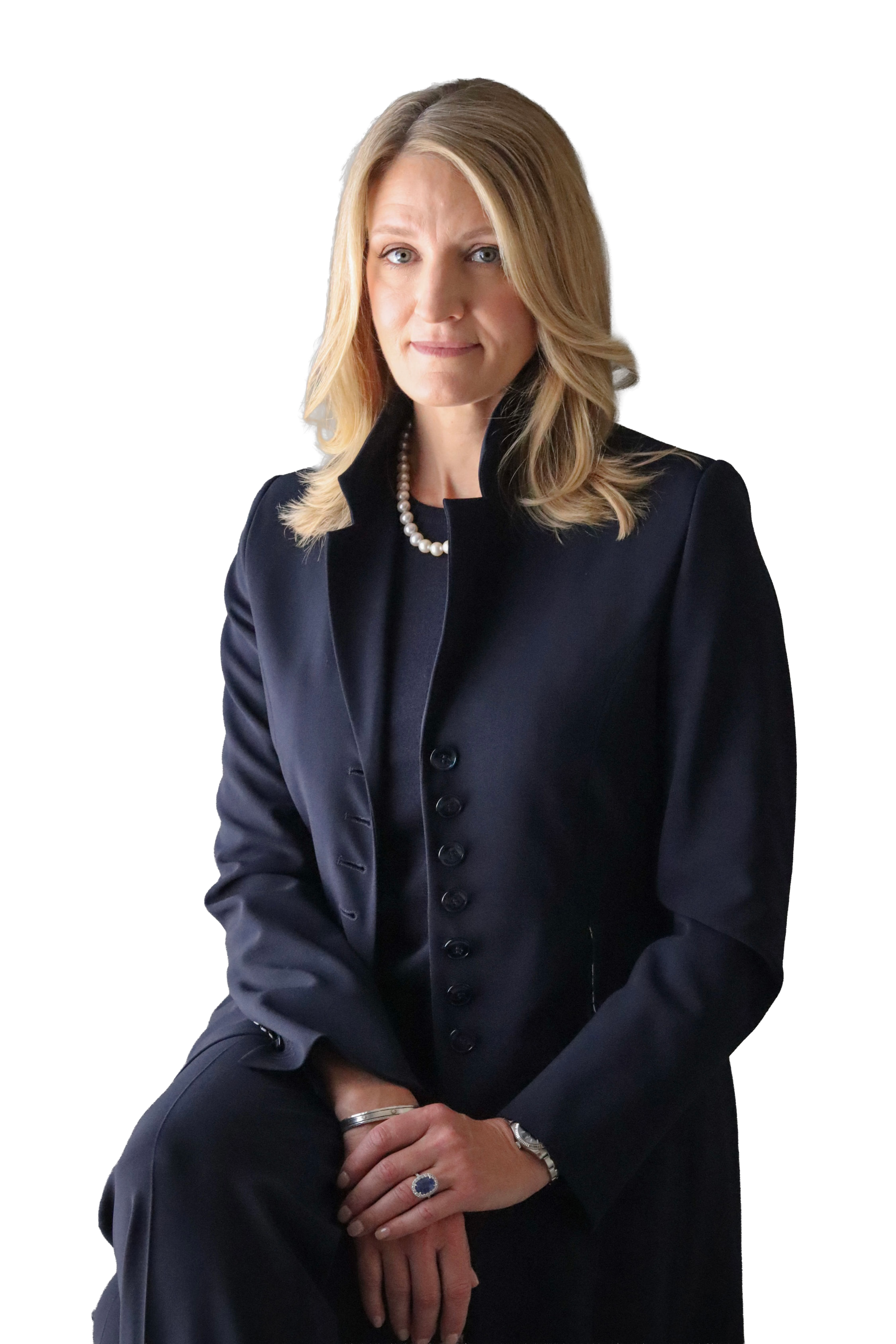 A woman with blonde hair styled in loose waves, wearing a navy blazer, a black top, a pearl necklace, and a ring, sitting with her hands clasped in her lap, against a plain background.