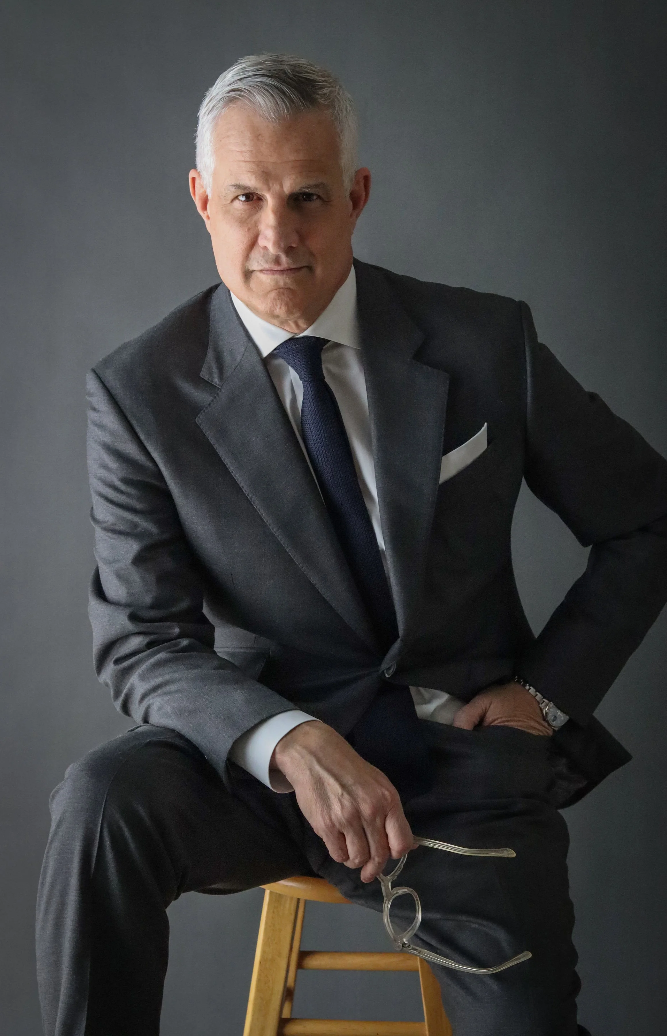 A man with gray hair in a suit sitting and holding glasses, looking down, with another person's arm in the foreground showing a watch and a white shirt cuff.