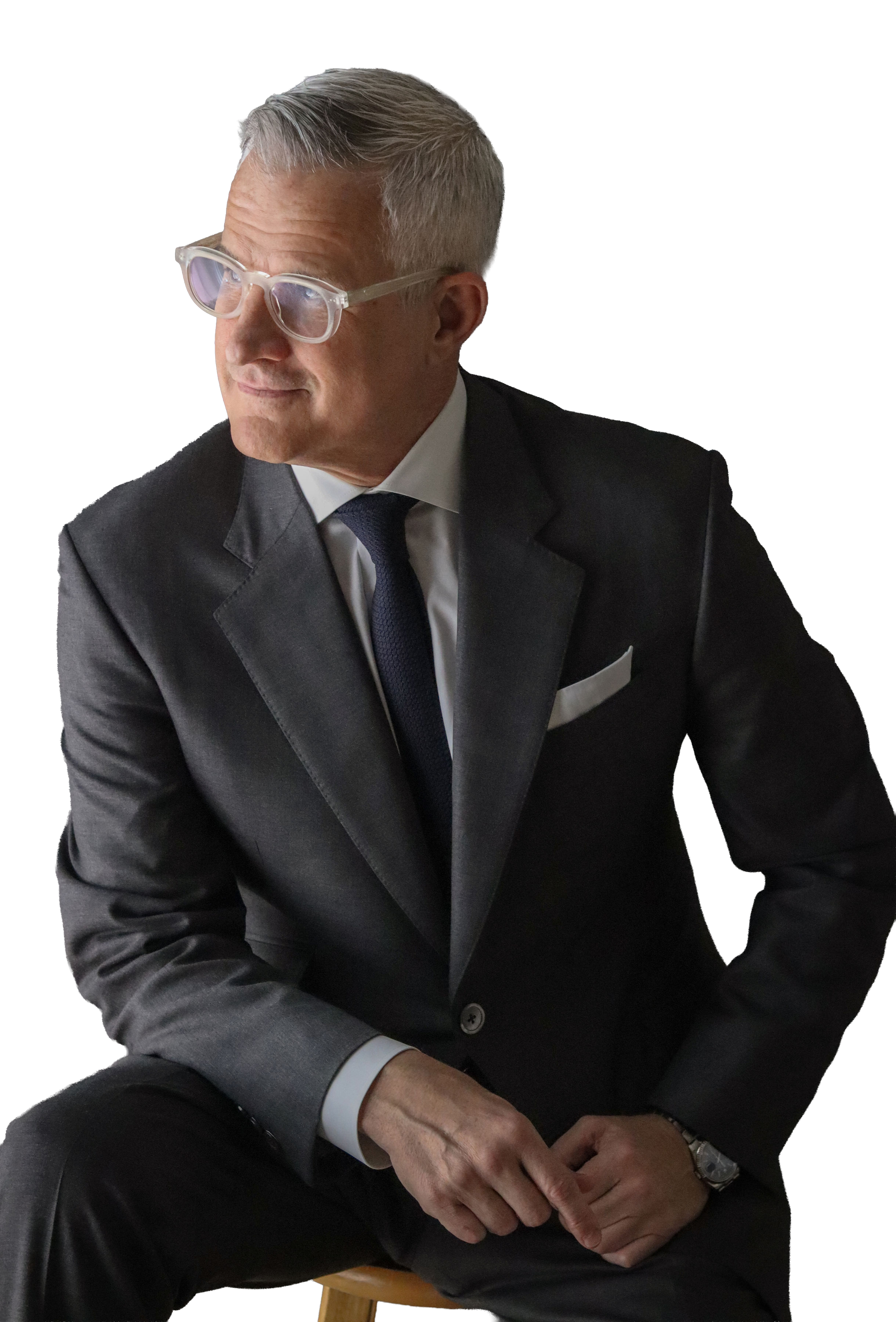 A man in a dark suit, white shirt, and dark tie sitting with his hands clasped, wearing glasses and a wristwatch, with a neutral expression.