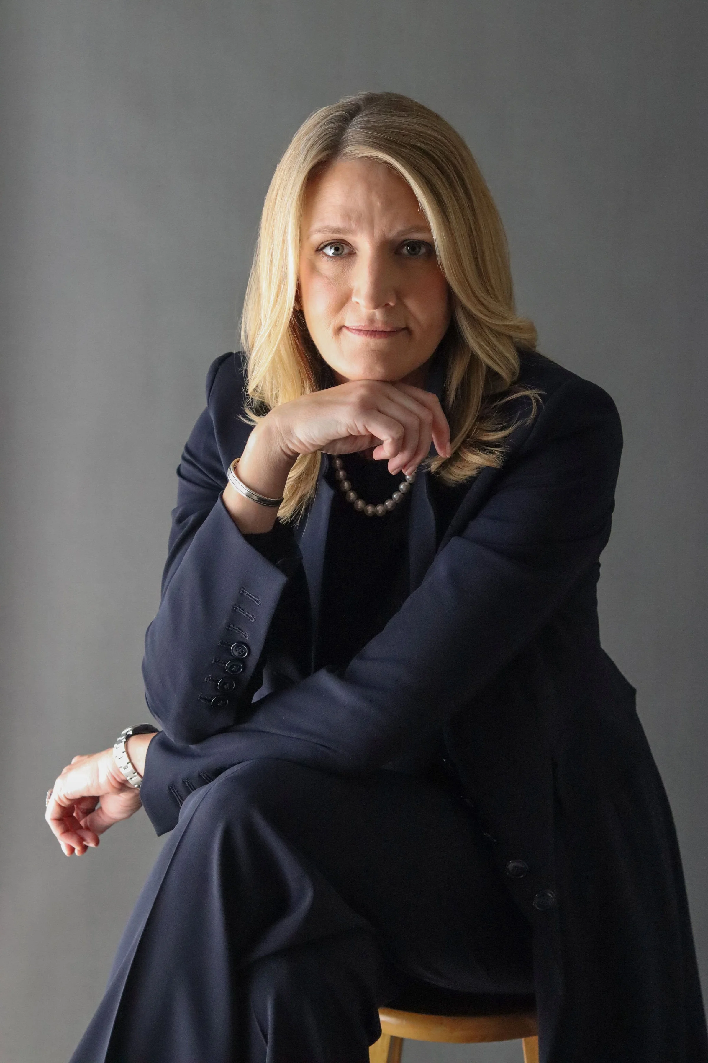 A woman with blonde hair wearing a dark suit, sitting on a chair with a gray background, resting her chin on her hand and looking at the camera.
