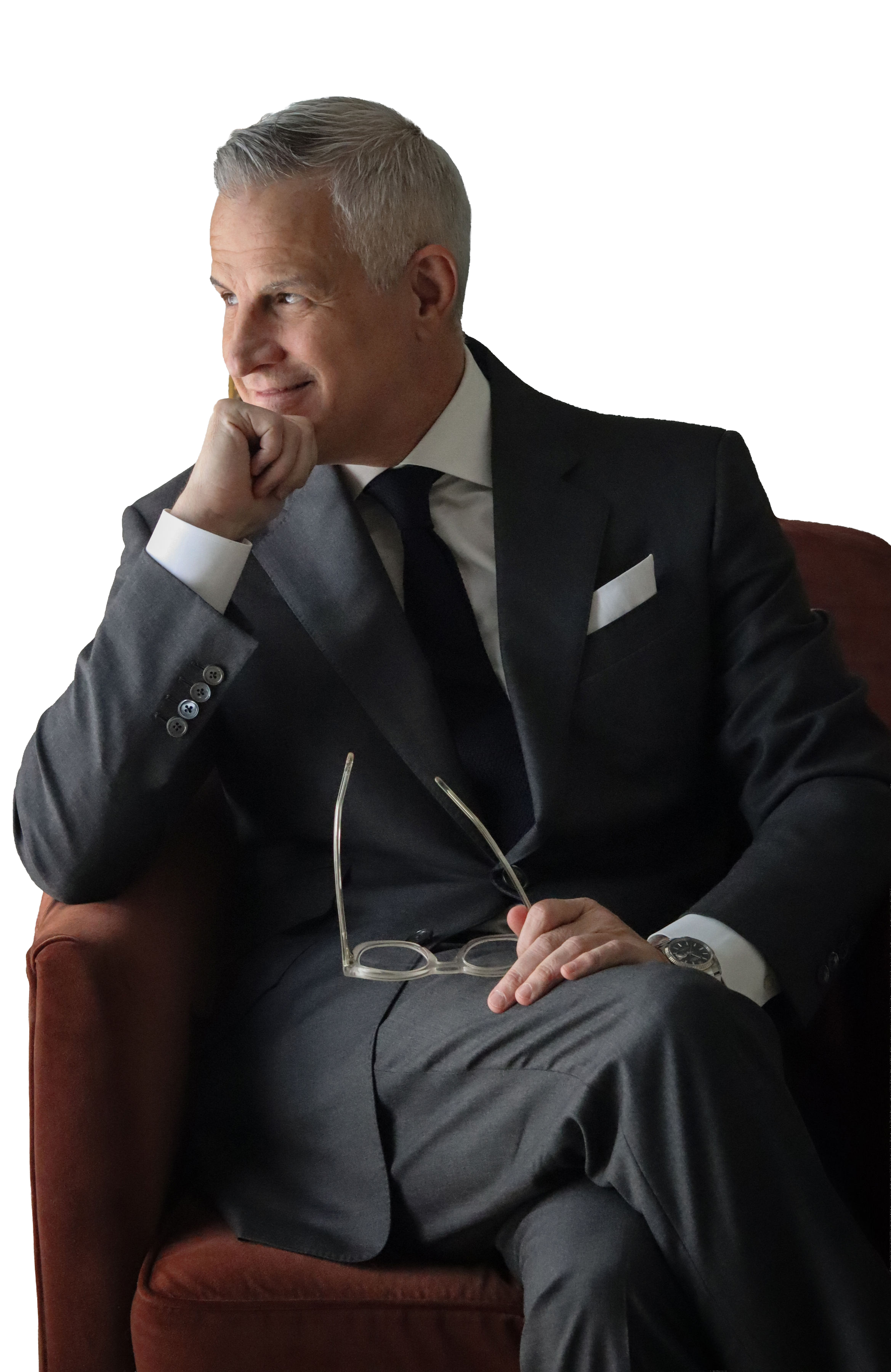 A man in a dark suit and white shirt sitting on a brown chair, holding reading glasses, with his chin resting on his hand, in a thoughtful pose.