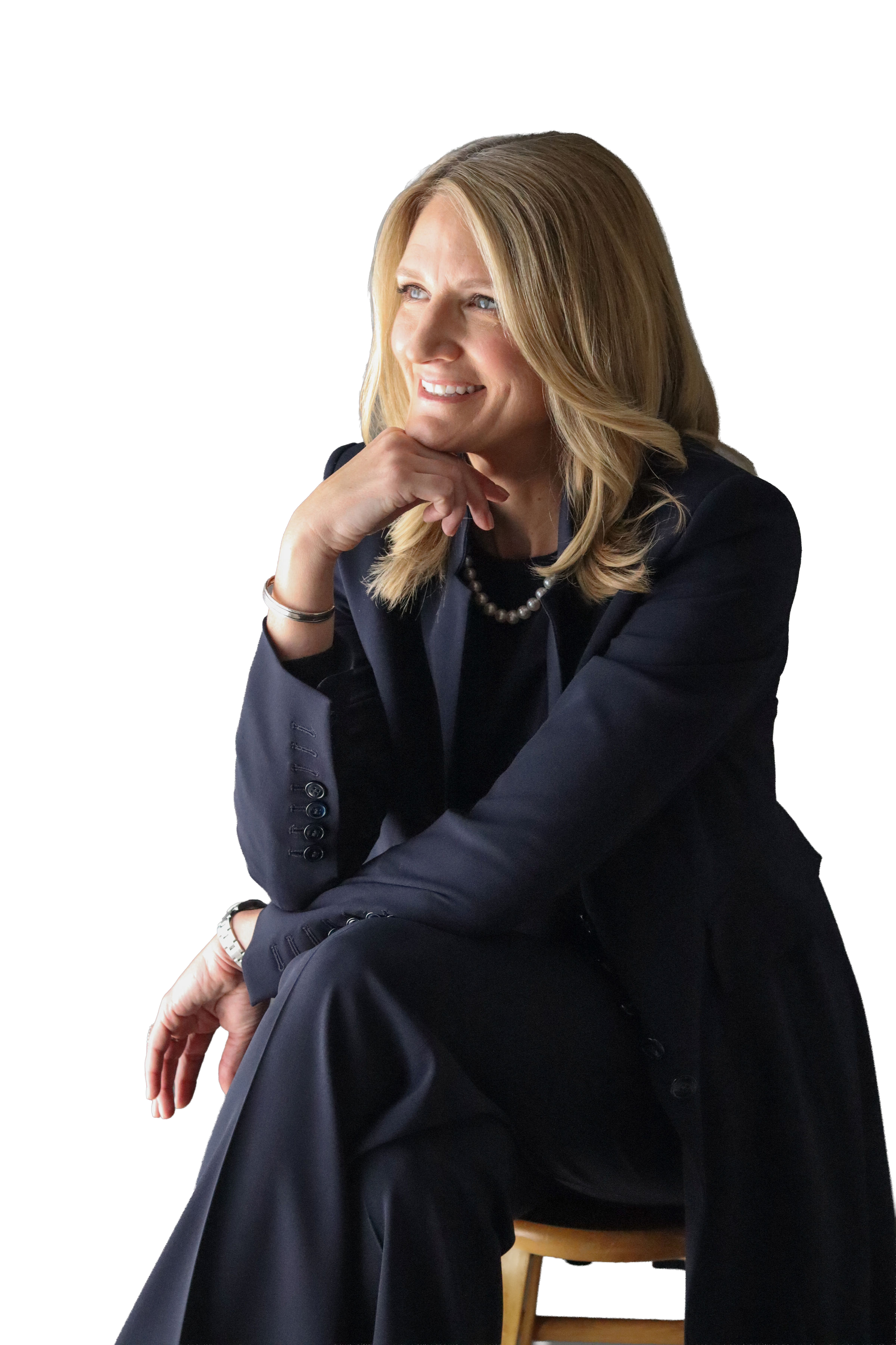 A woman with blonde hair, wearing a dark suit and pearl necklace, sitting on a stool with her chin resting on her hand, smiling thoughtfully.