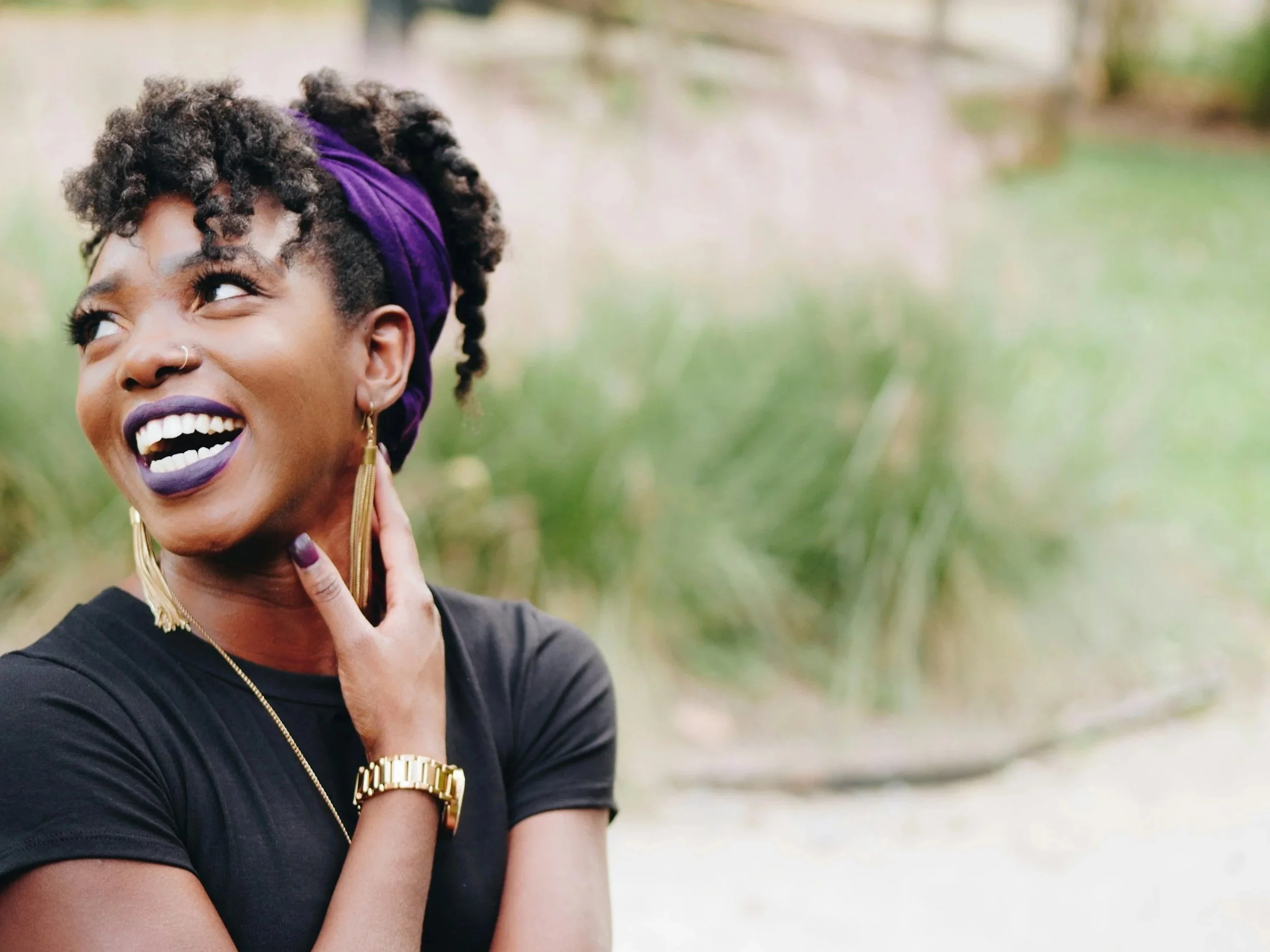 Close-up of a smiling woman with dark curly hair styled up and wrapped with a purple headscarf, wearing makeup including purple lipstick, gold earrings, and a gold watch, outdoors with blurred green background.