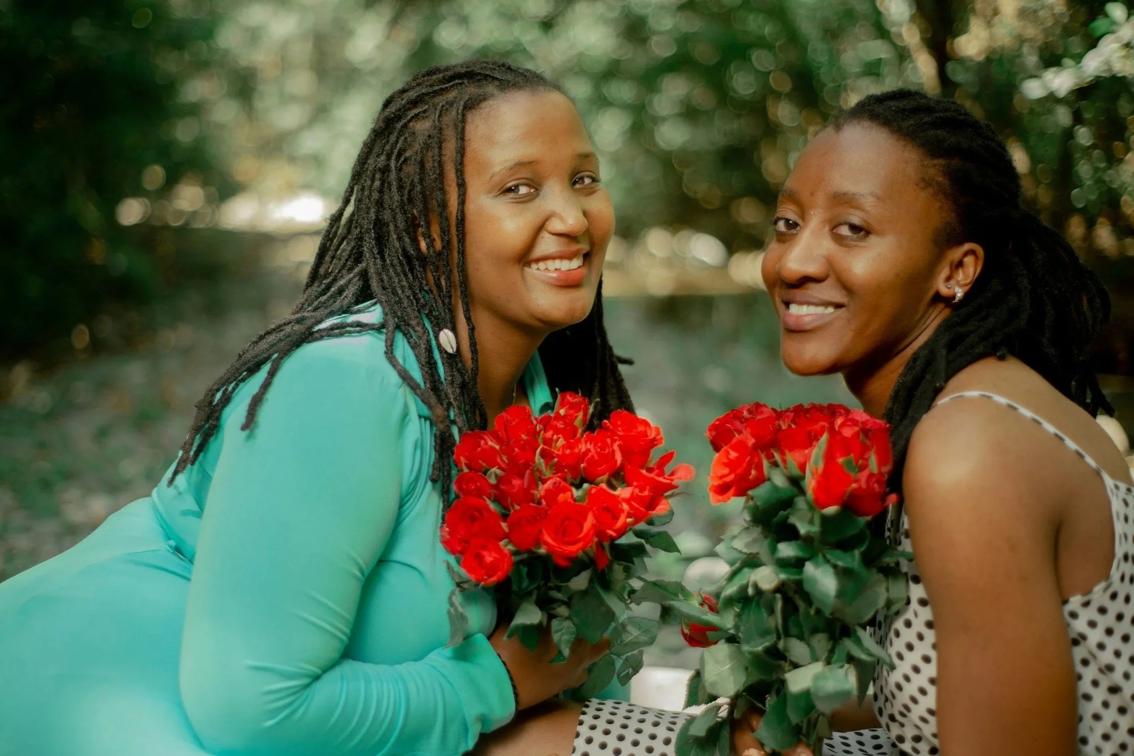 Two women smiling and holding bouquets of red flowers, outdoors with a blurred green background.
