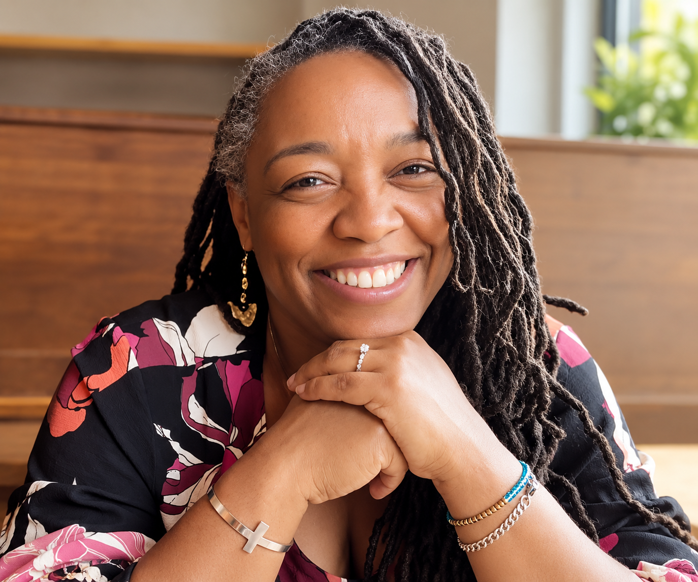 A smiling Black woman with dreadlocks, wearing earrings and jewelry, resting her chin on her hands, indoors with a wooden background and window with natural light.