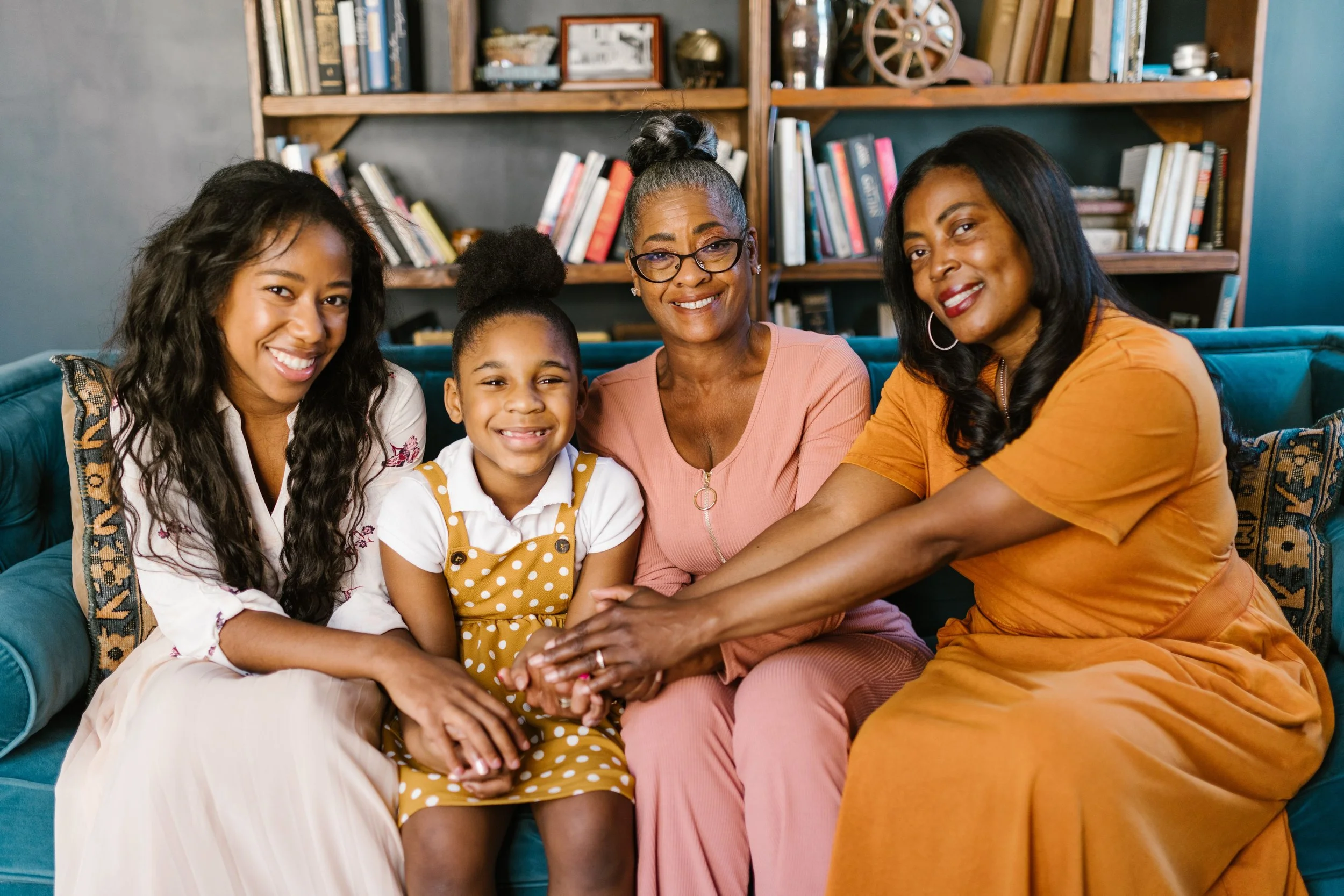 A multigenerational African American family sitting on a sofa, smiling, with a bookshelf in the background.