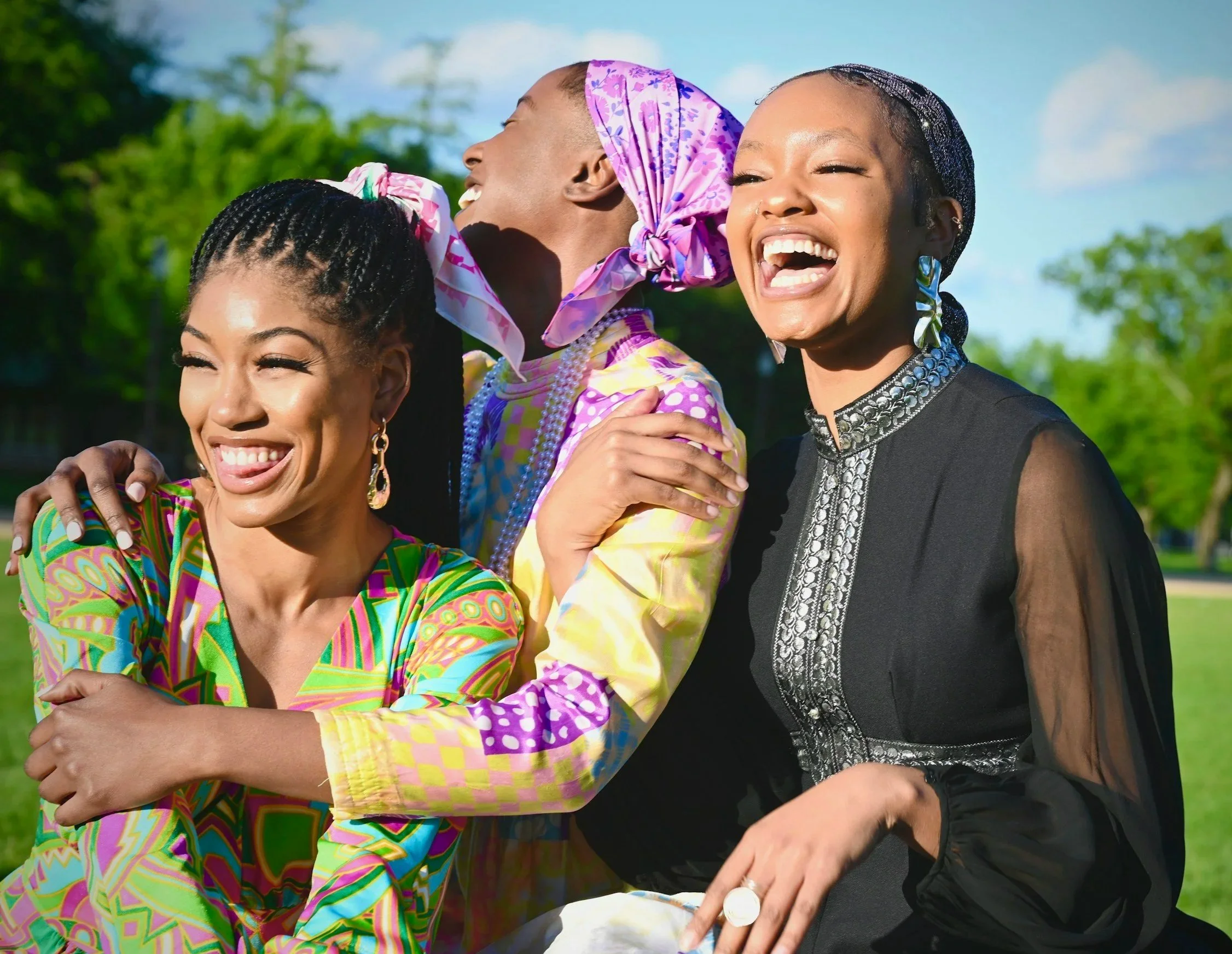 Three women smiling and embracing outdoors against a background of trees and a blue sky.