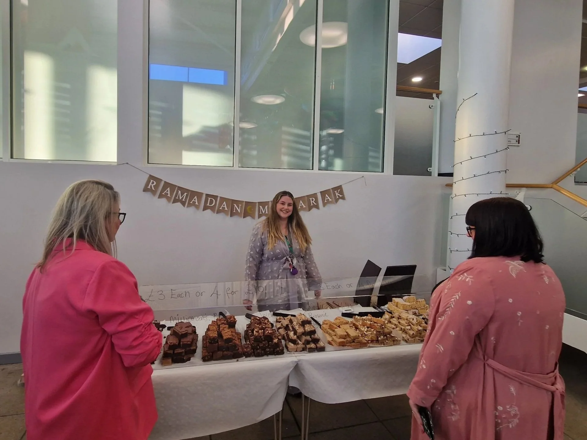 The Fabled Bakery owner at a dessert stand with brownies, with a woman smiling behind the counter. A banner above reads "Ramadan Marak," and there are handwritten signs and a variety of baked goods on display.