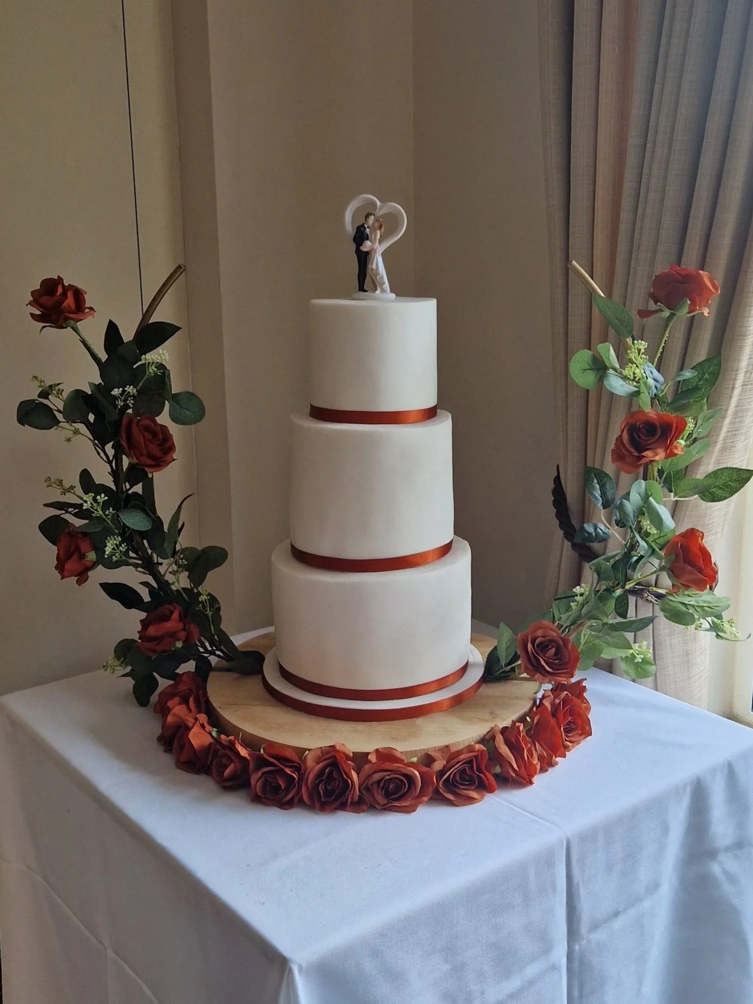 A three-tier wedding cake decorated with a bride and groom topper, surrounded by red roses and greenery, on a wooden base with a white tablecloth.