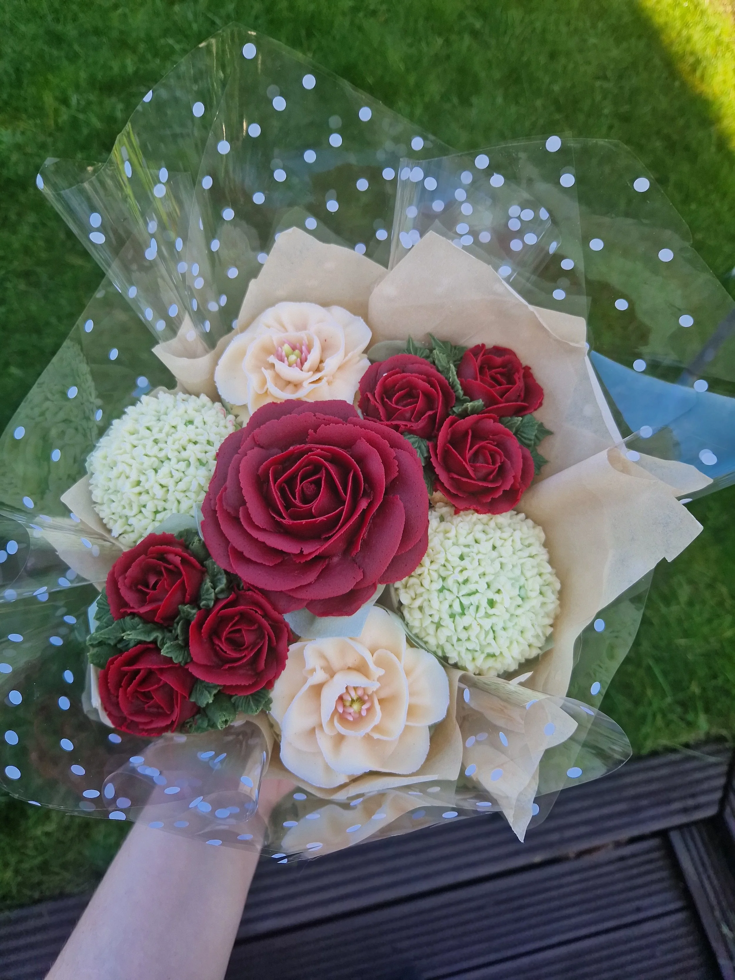 A cupcake bouquet of red and cream-colored flowers wrapped in clear polka-dotted plastic on a green grassy background.
